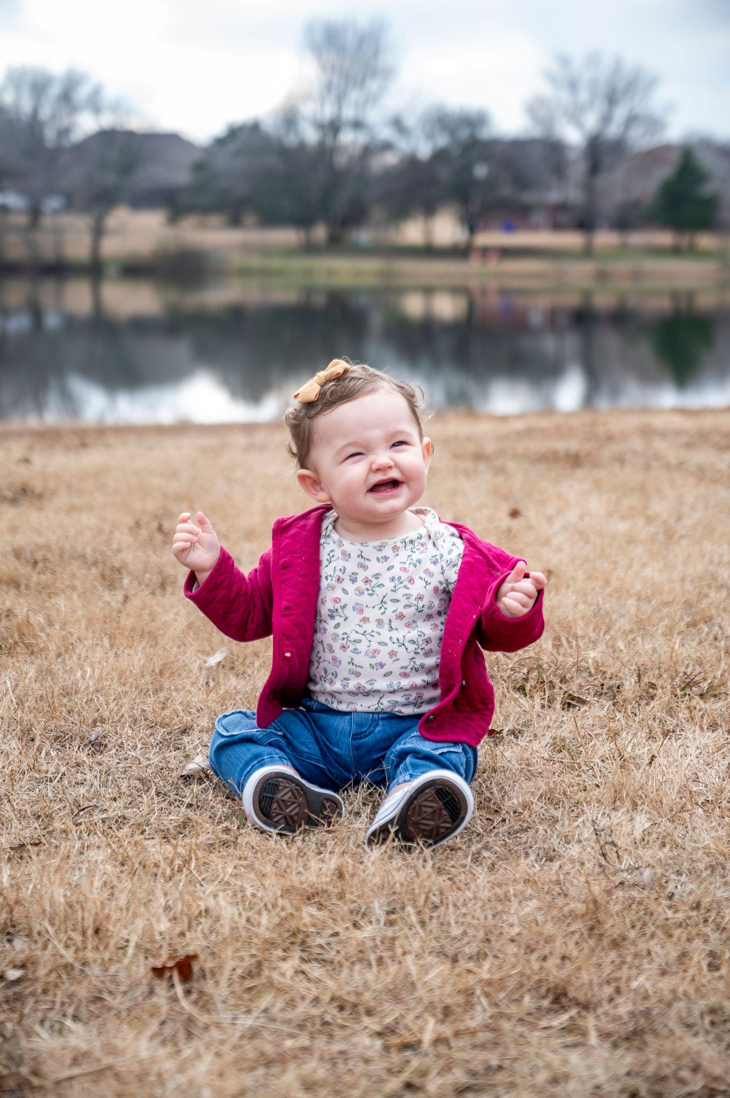 A happy baby in a pink cardigan sits on brown grass near a lake.