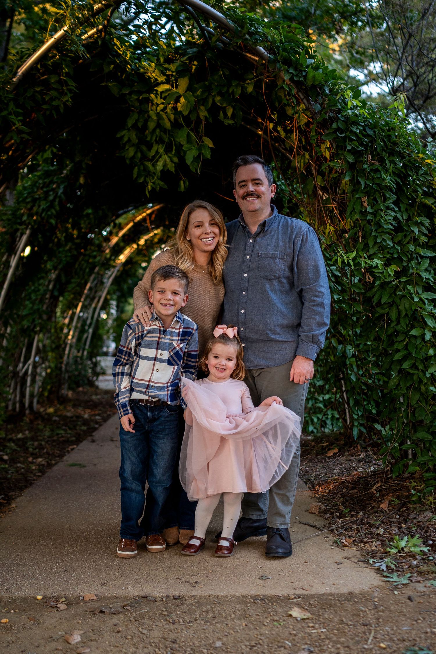 A happy family of four poses under a lush green garden archway, smiling together for a portrait photo.