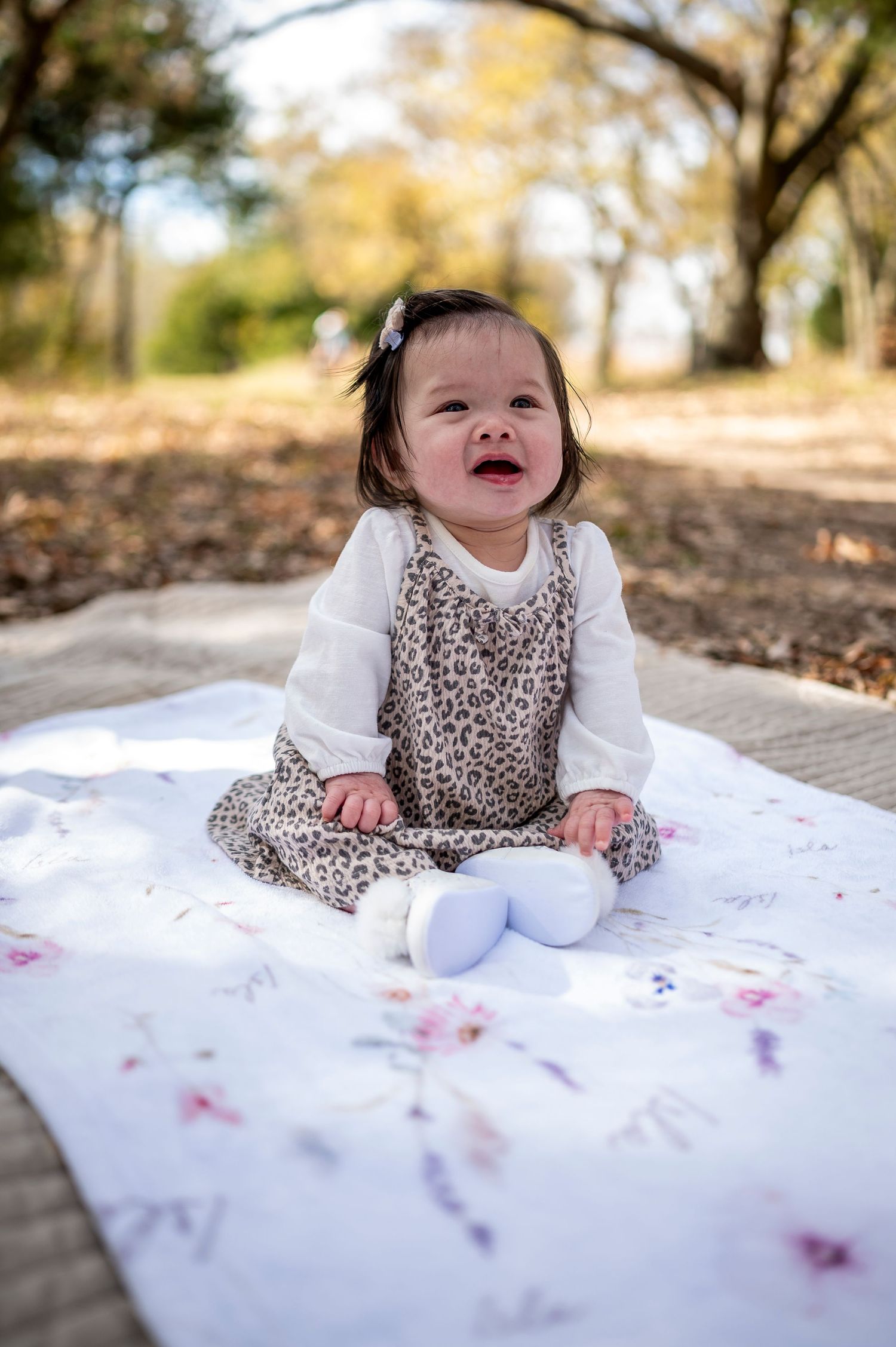 Adorable baby girl in leopard print romper sits on floral blanket outdoors, smiling in natural park setting.