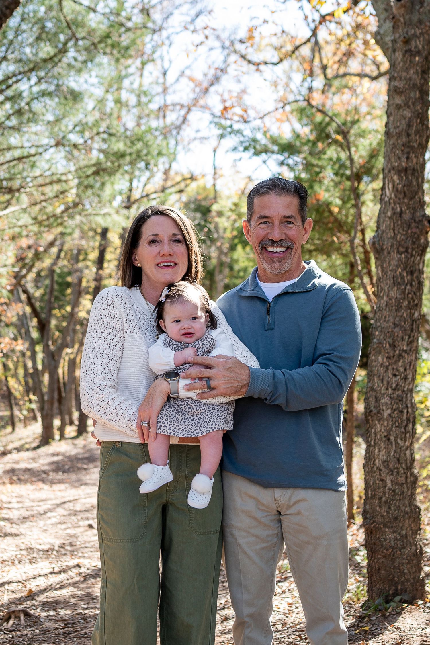 Smiling couple holding baby girl in floral dress outdoors among autumn trees in a family portrait.