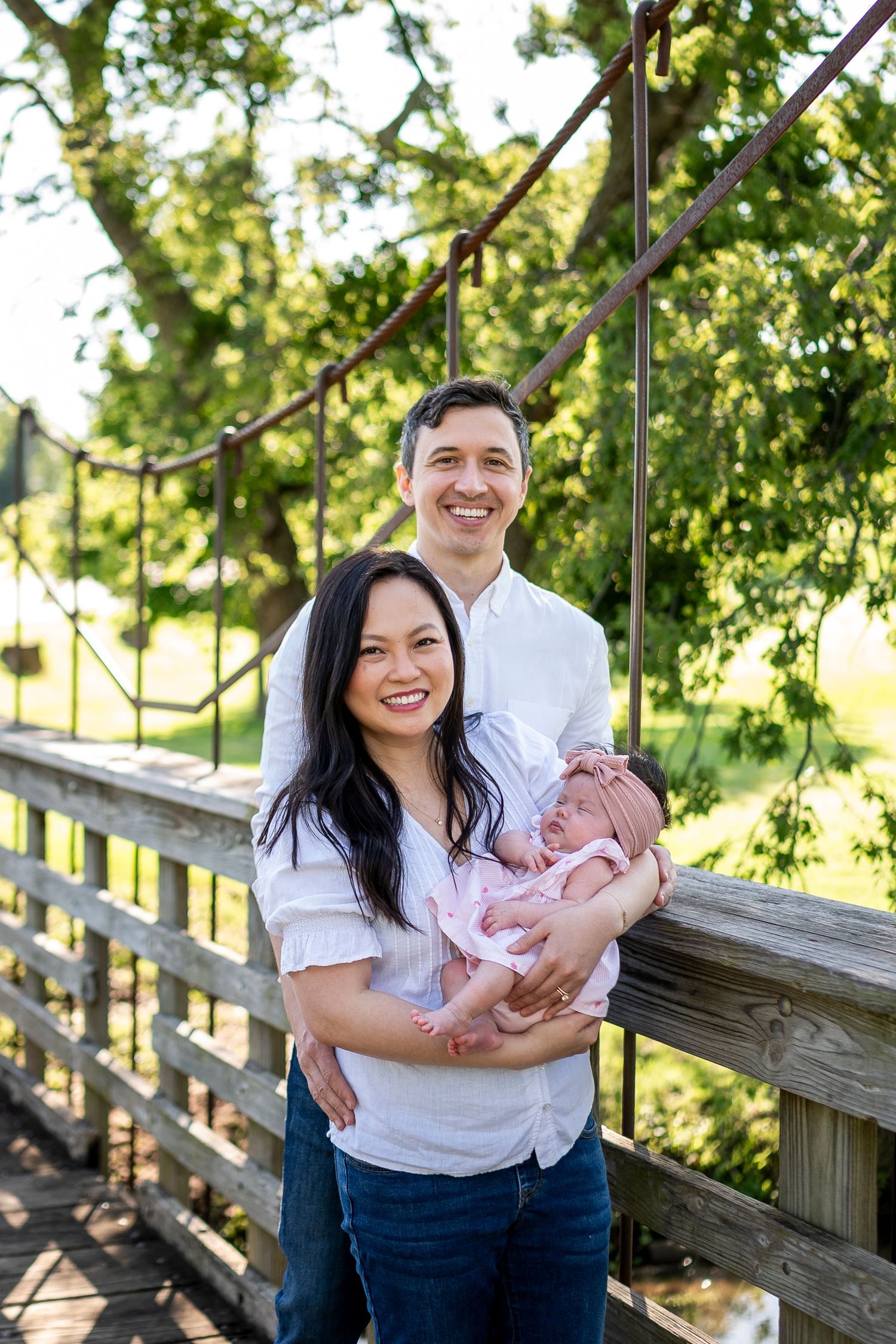 A family sits together on a wooden bridge surrounded by lush green trees while holding a small baby.