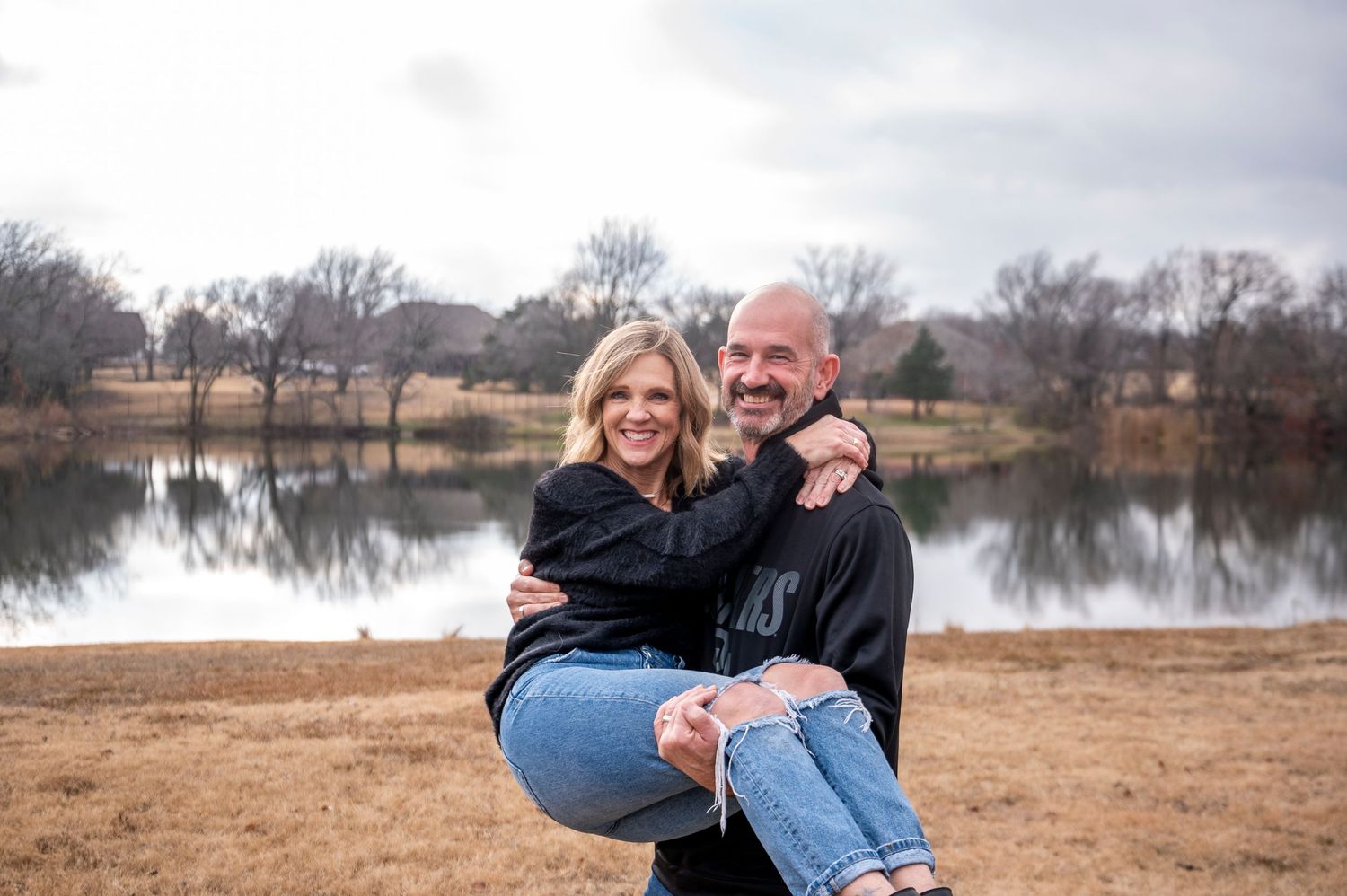 Couple sharing playful moment by lake with cloudy winter sky.