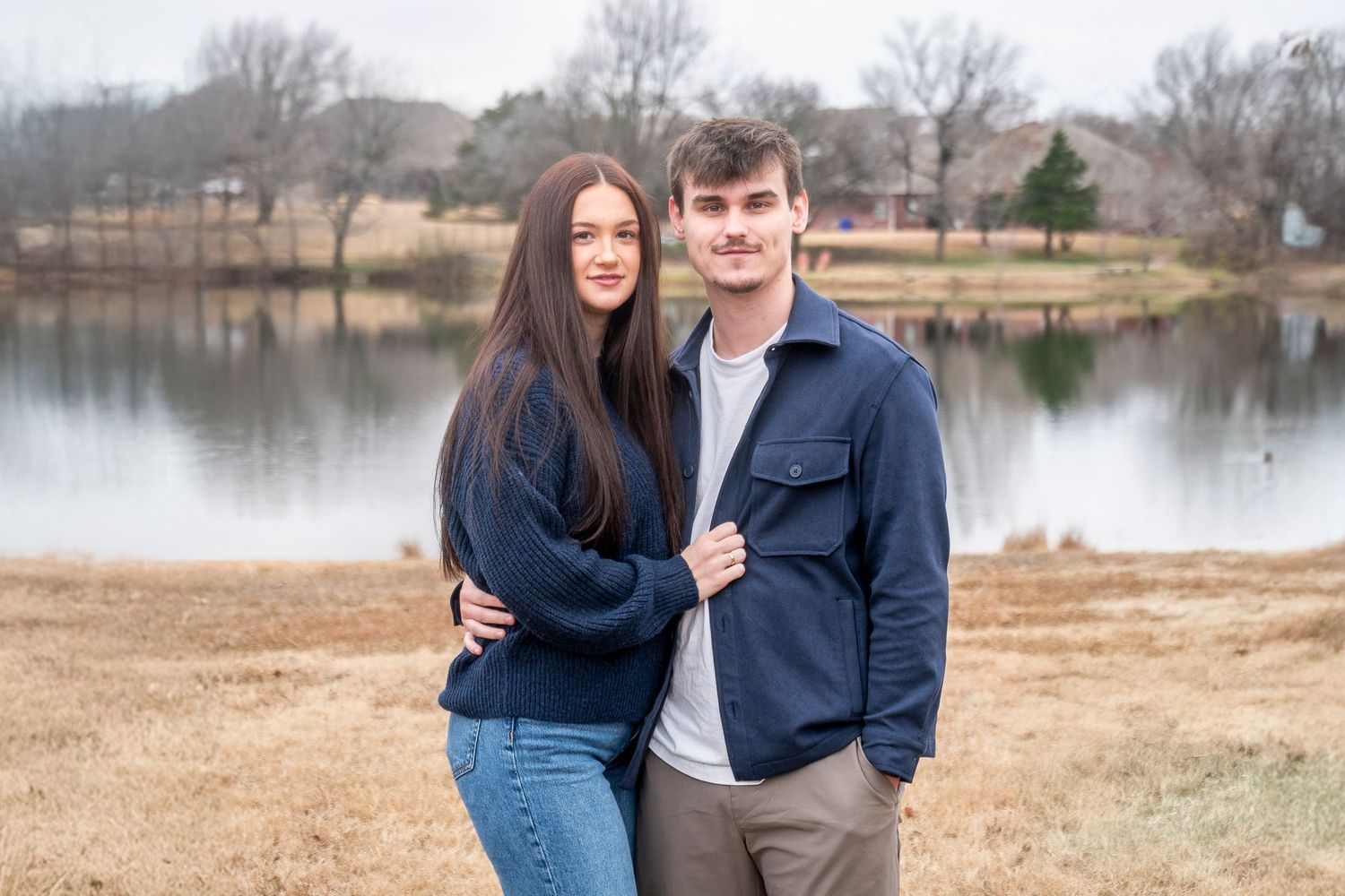 Couple in casual outfits embracing by a peaceful lakeside landscape.