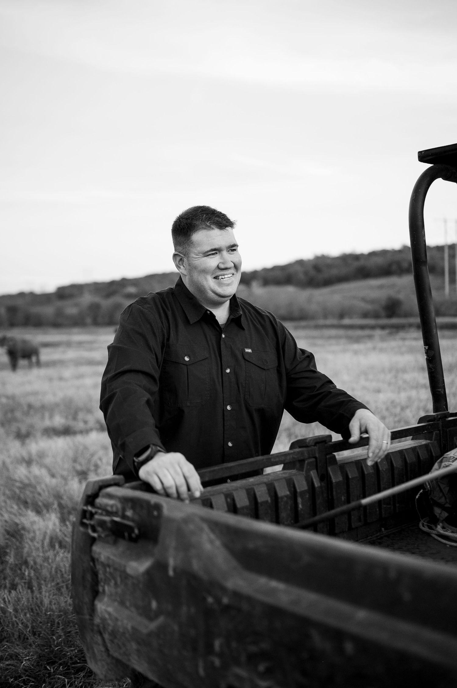 Black and white photo of smiling man leaning on farm equipment in a rural field setting.