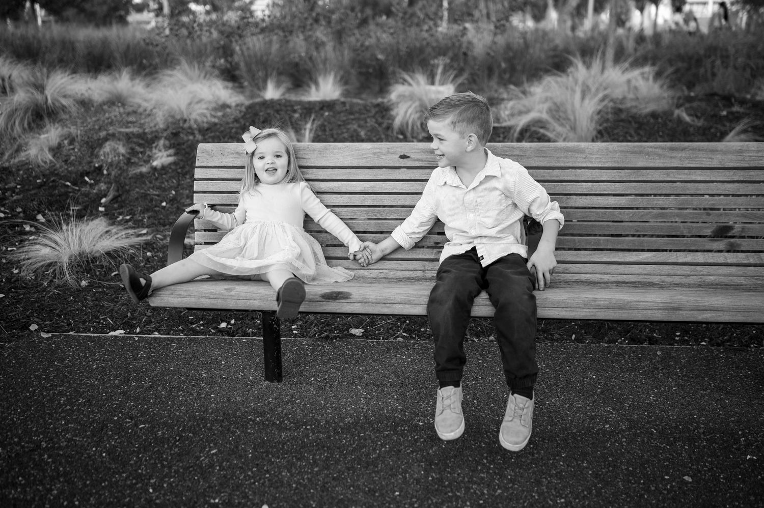 Two children sit and interact playfully on a wooden bench surrounded by ornamental grasses.