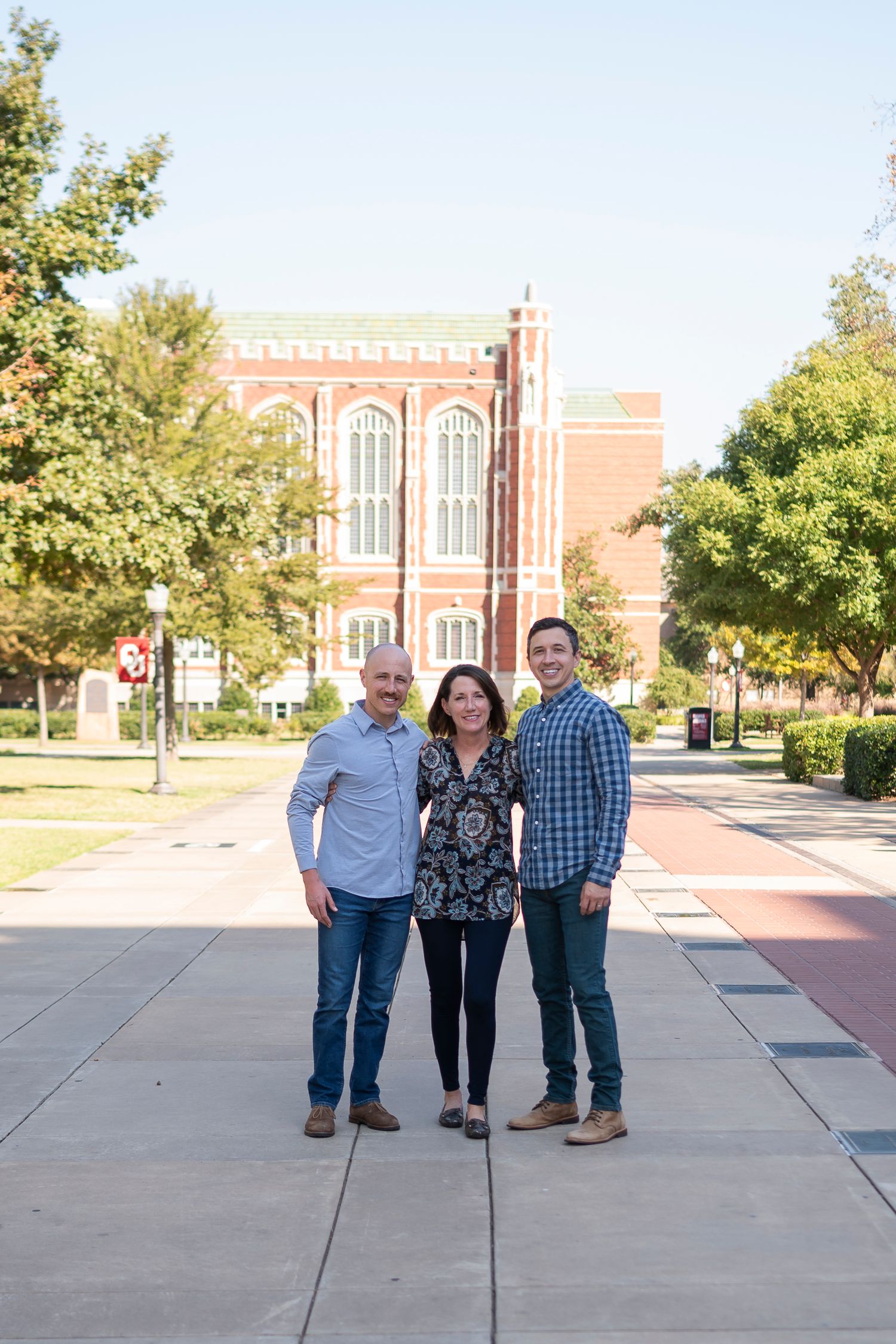 Three people standing together on campus walkway with historic brick building in background.