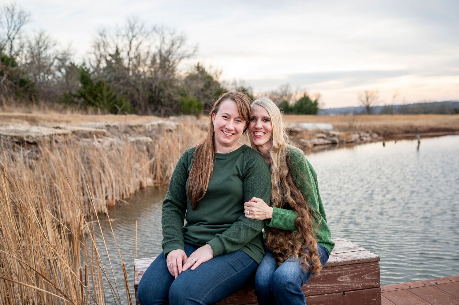 Two smiling women in matching green sweatshirts sit on a dock by a calm lake surrounded by tall grass at sunset.