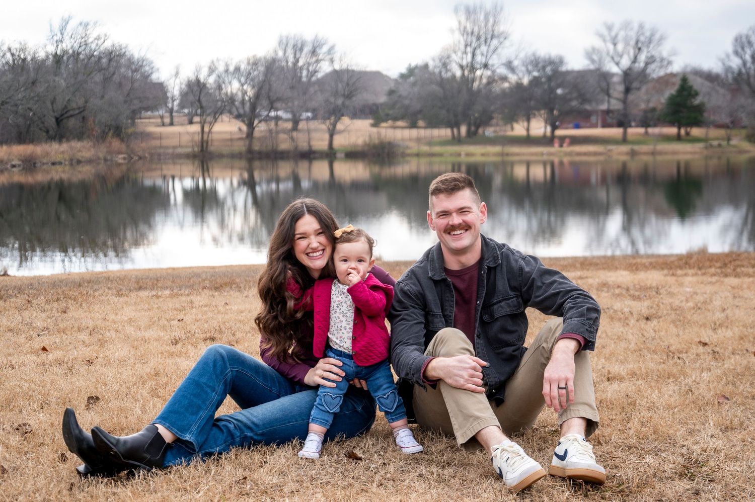 Family of three sitting together on brown grass by a winter lake.
