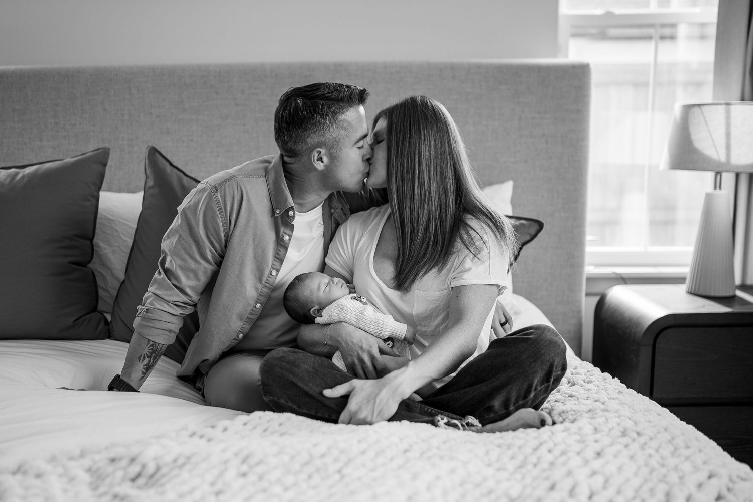 Black and white photo of a couple kissing on a bed while holding their newborn baby.