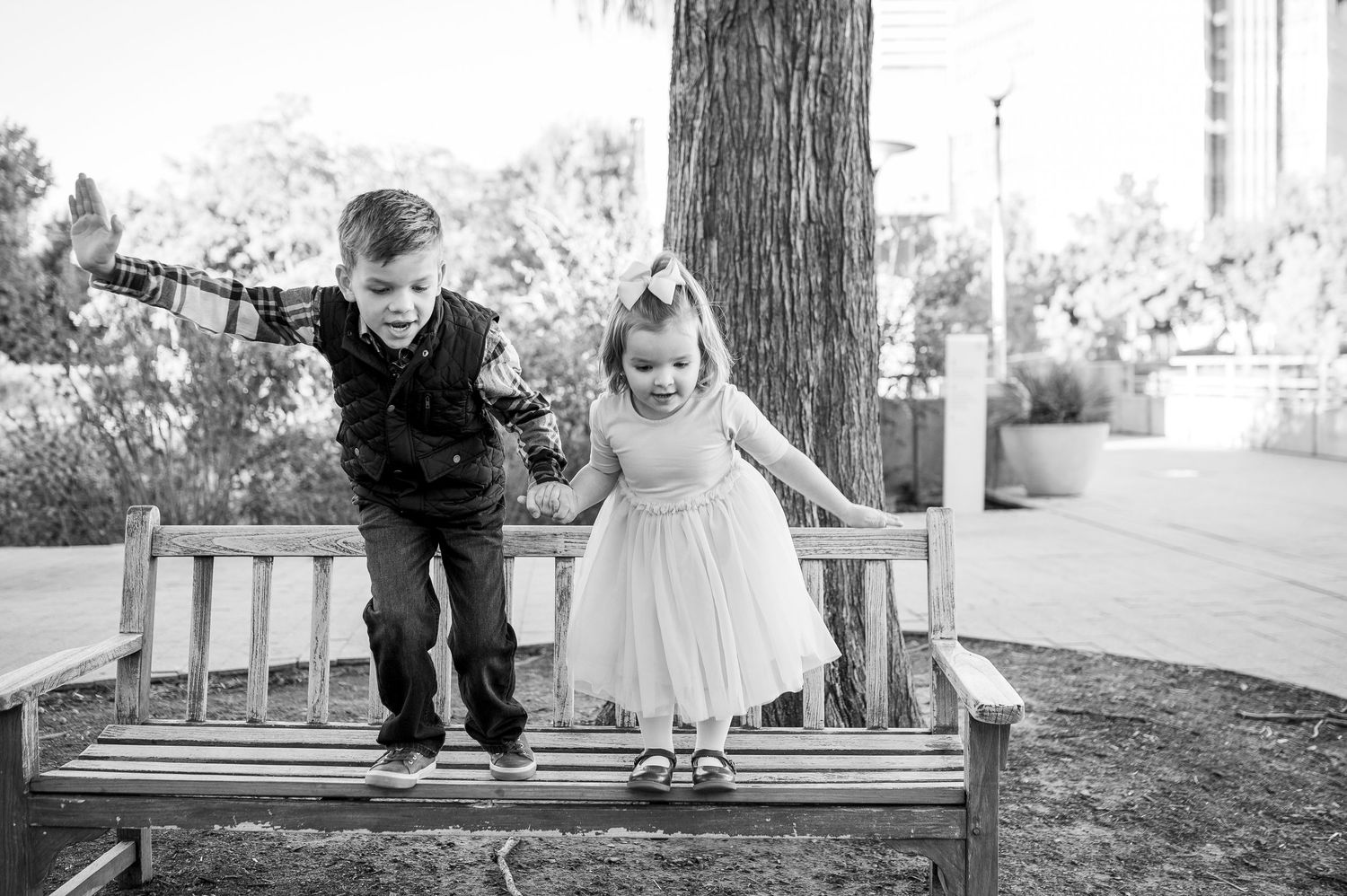 Two young children in formal attire playfully stand on a wooden bench outdoors in a black and white photo.