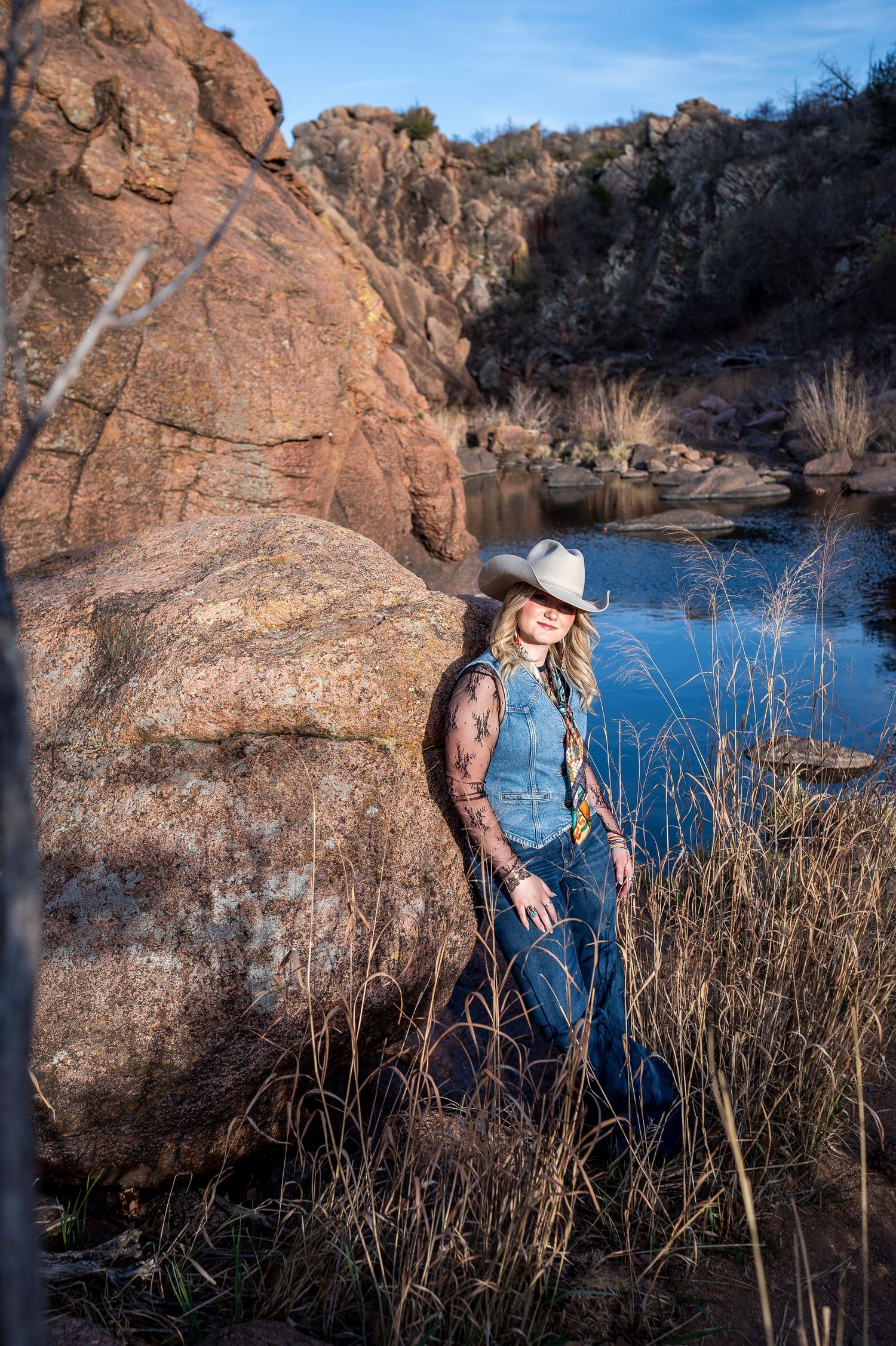 Blonde woman in white cowboy hat and denim vest leans against boulder near a rocky river in natural landscape.