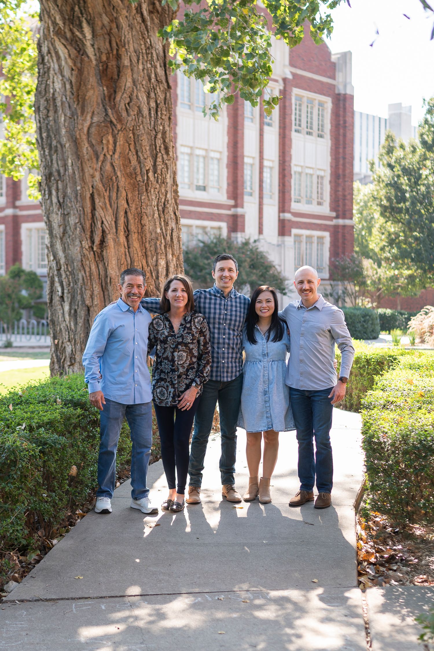 Group photo on campus pathway with large tree trunk and red brick university building in background.