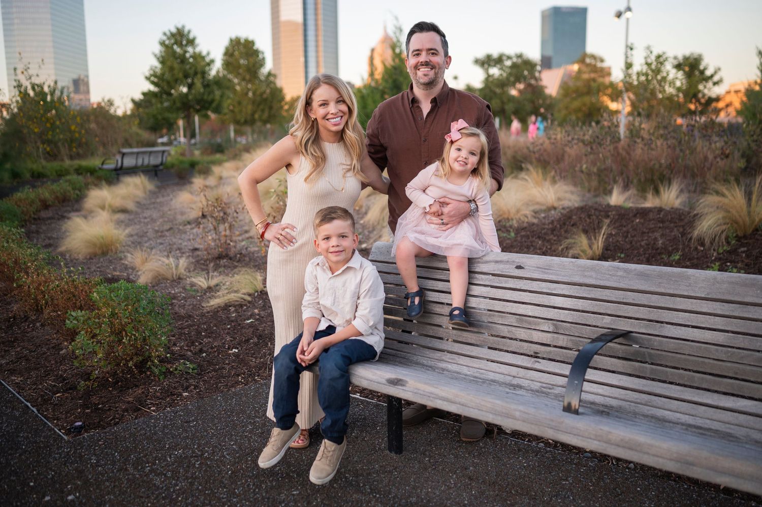 A family of four sits together on a wooden bench with urban architecture in the background.