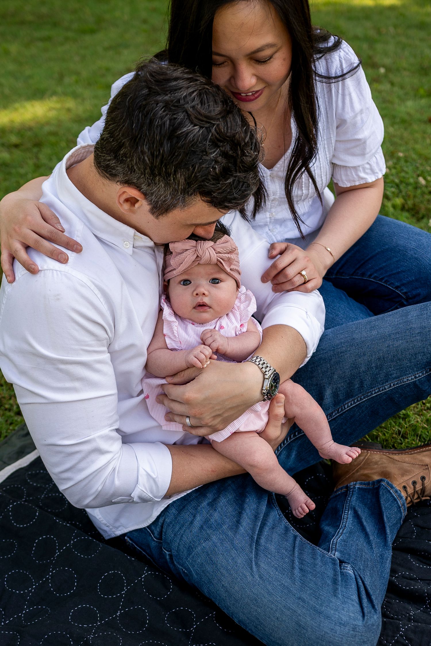 Parents in white shirts and jeans share tender moments with their baby on a sunny lawn.
