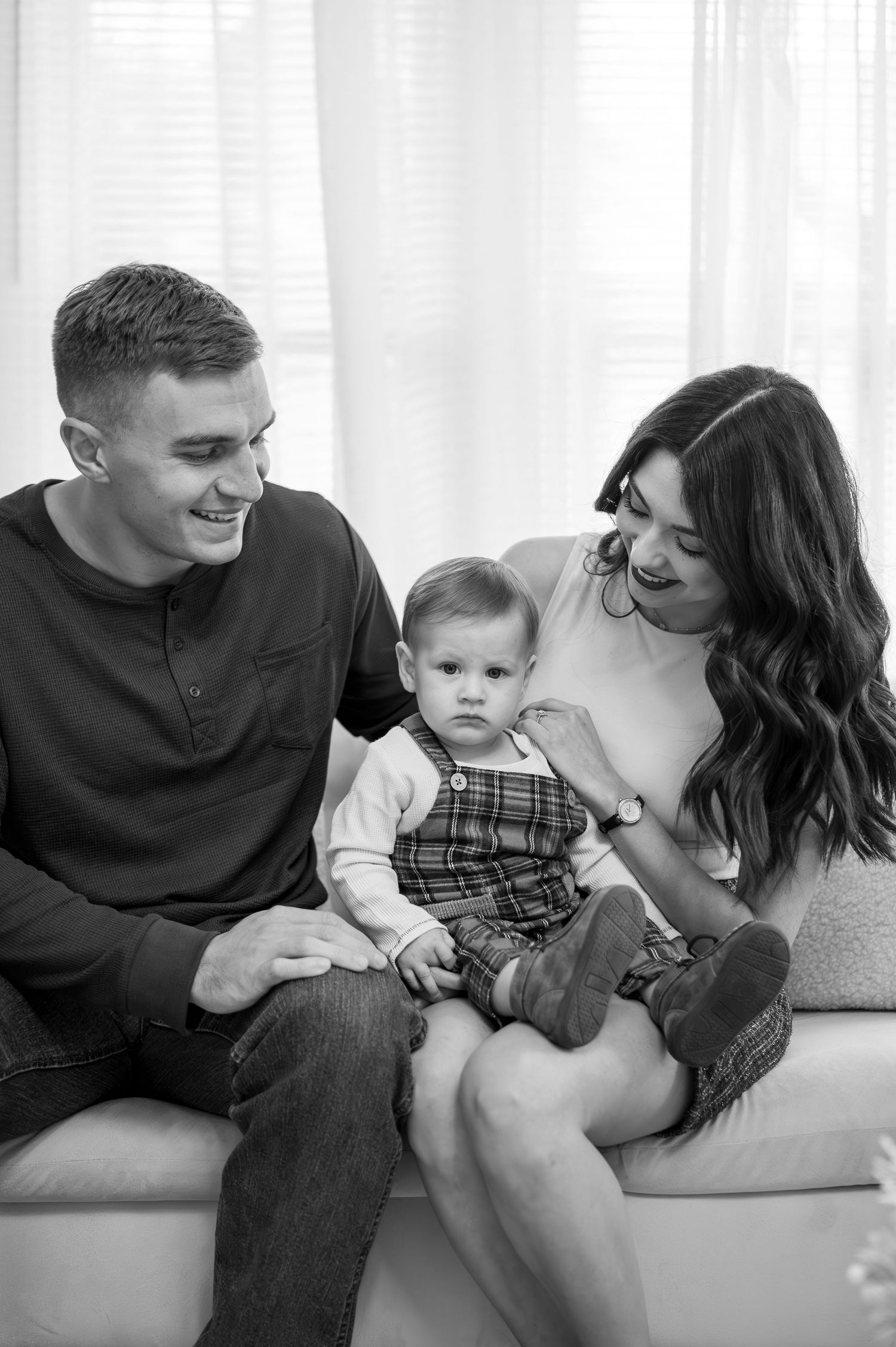 A young couple sits on a couch with their baby wearing plaid overalls in a candid black and white portrait.