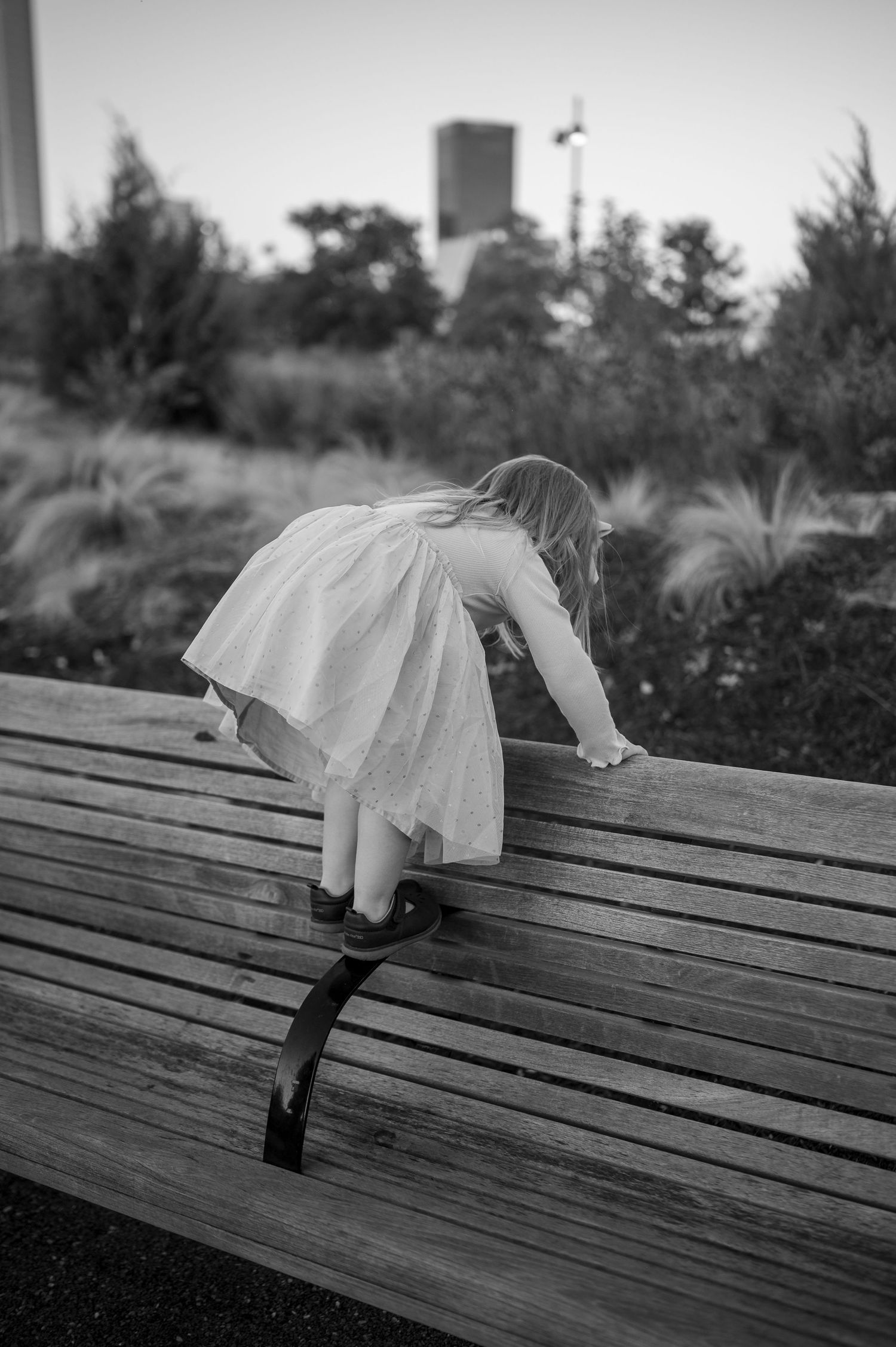 A child in a white dress climbing on a wooden bench in black and white photography.