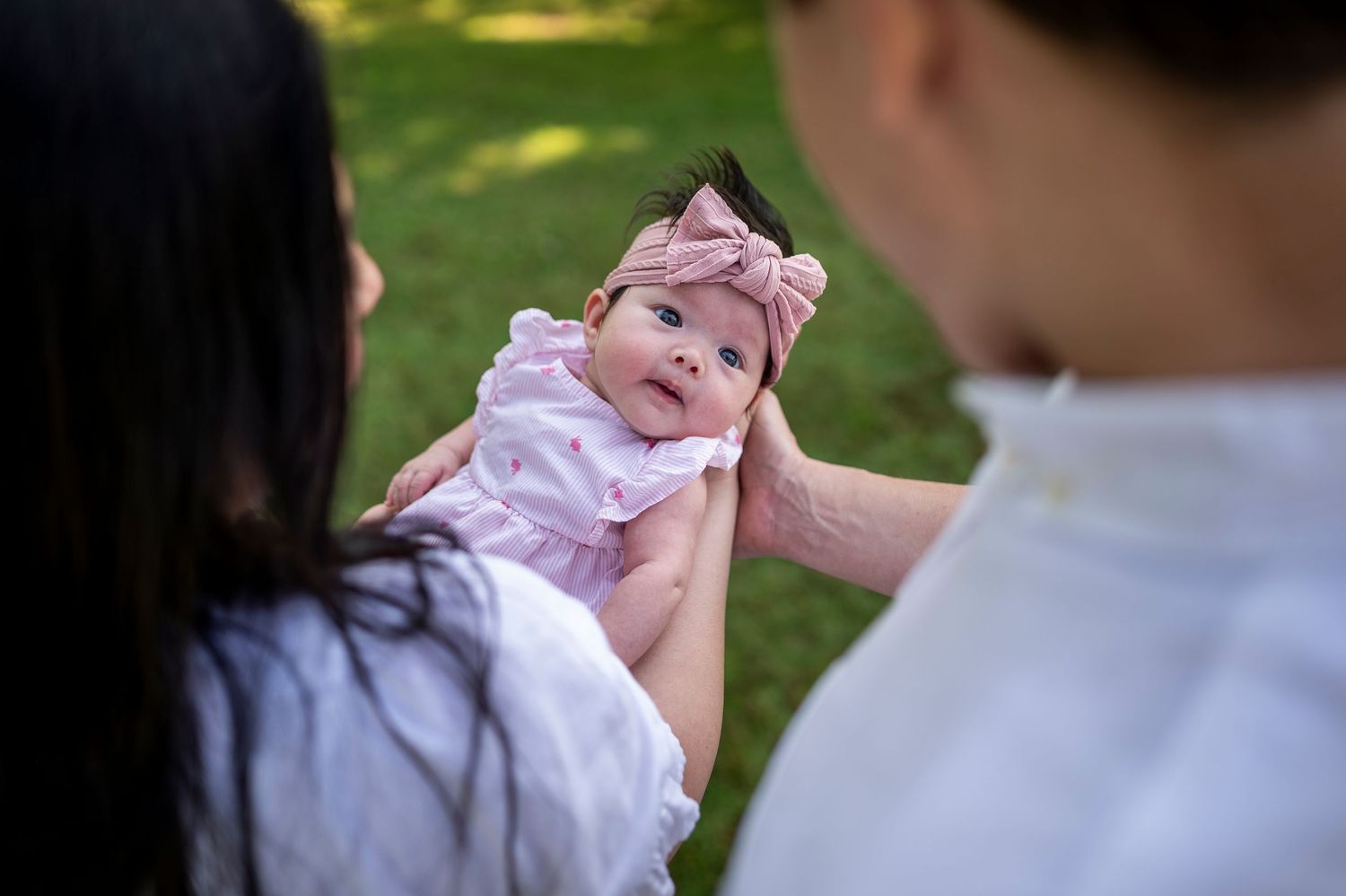 A baby wearing a pink headband and outfit is held up outdoors in a sunny garden setting.