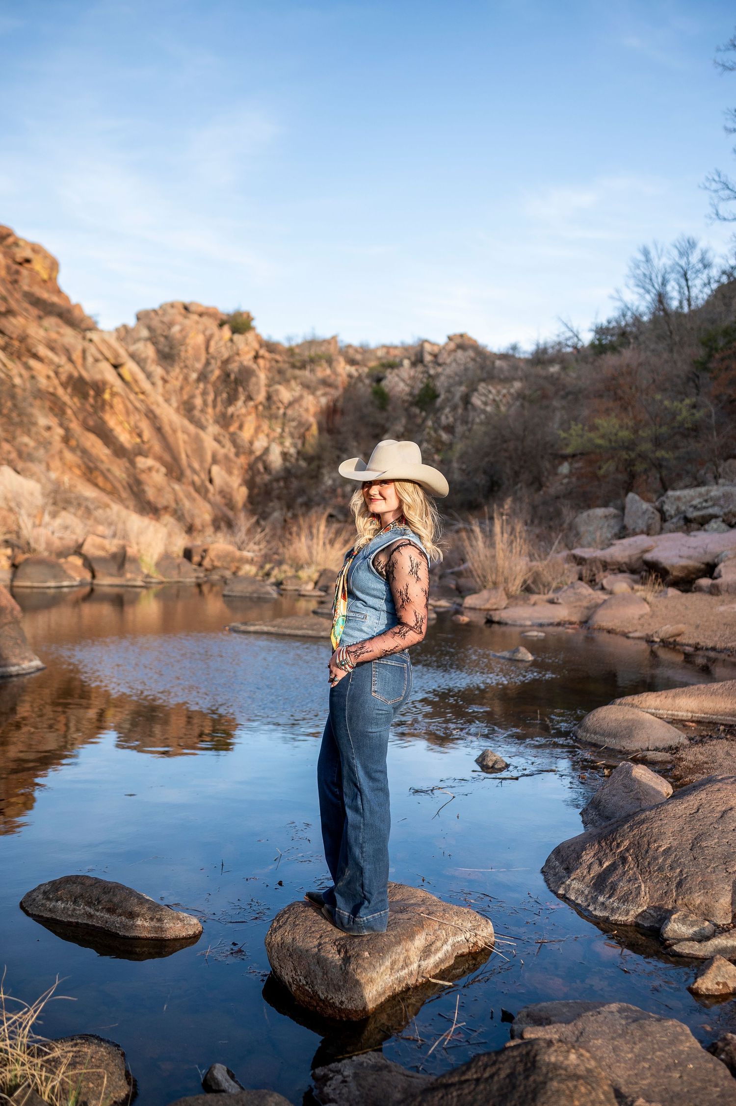 Woman in cowboy hat and denim overalls standing on rocks by a serene river with red rock canyon backdrop.
