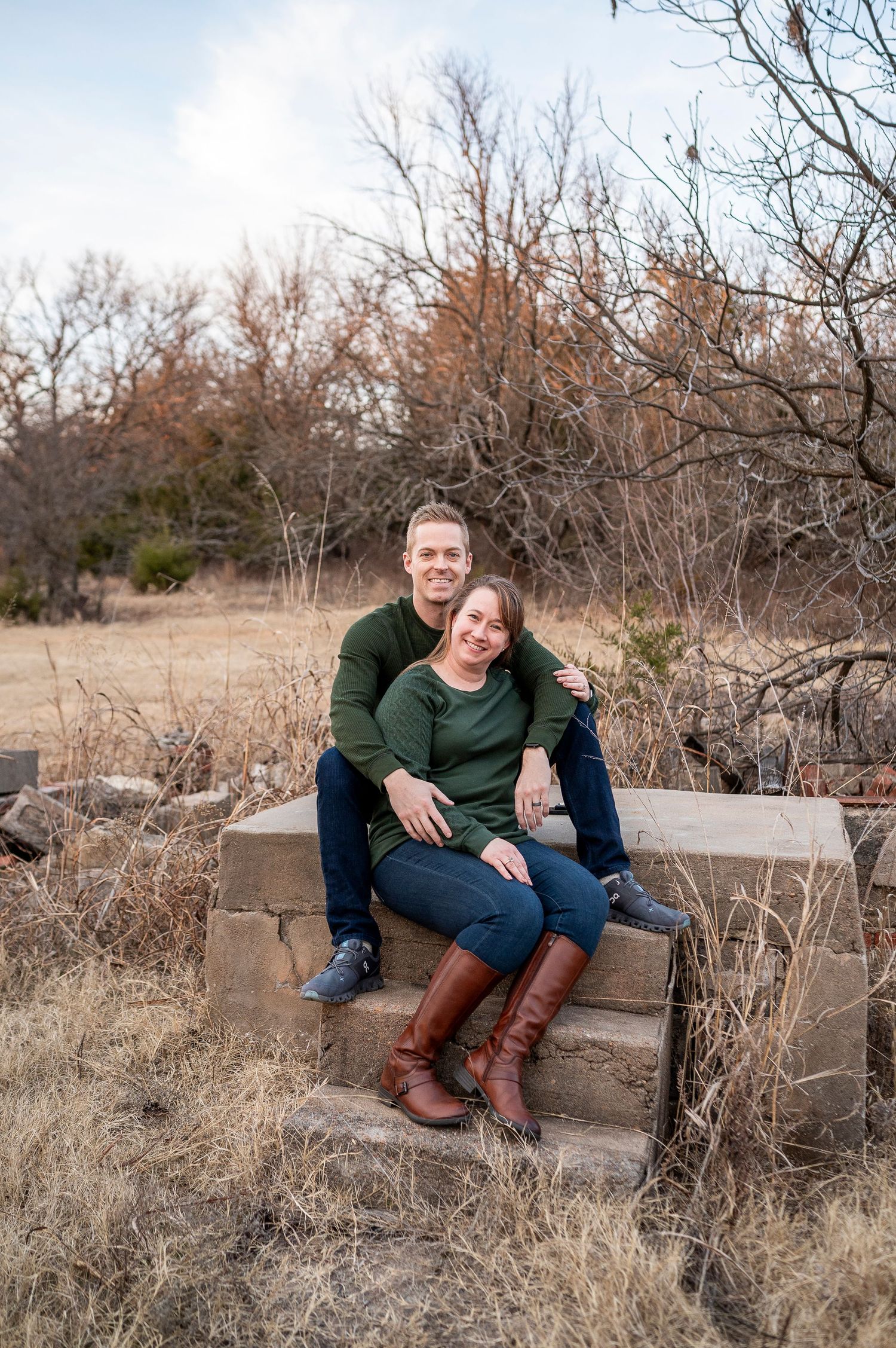 Couple in matching green tops and jeans sitting on stone steps in a rustic outdoor setting, smiling warmly.