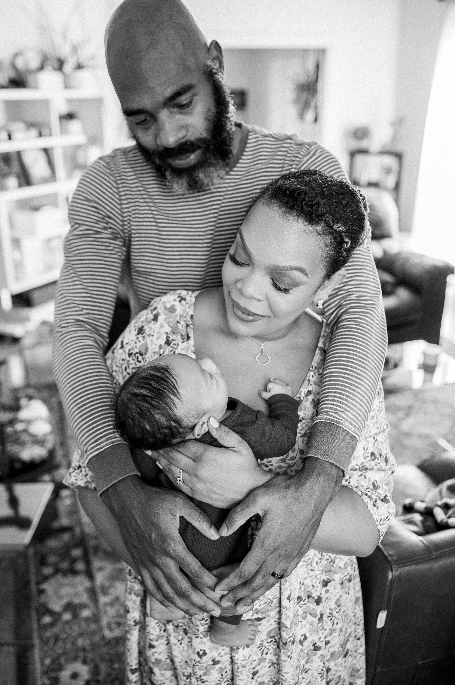 Black and white photo of a couple lovingly cradling their newborn baby, forming a heart shape with their hands.