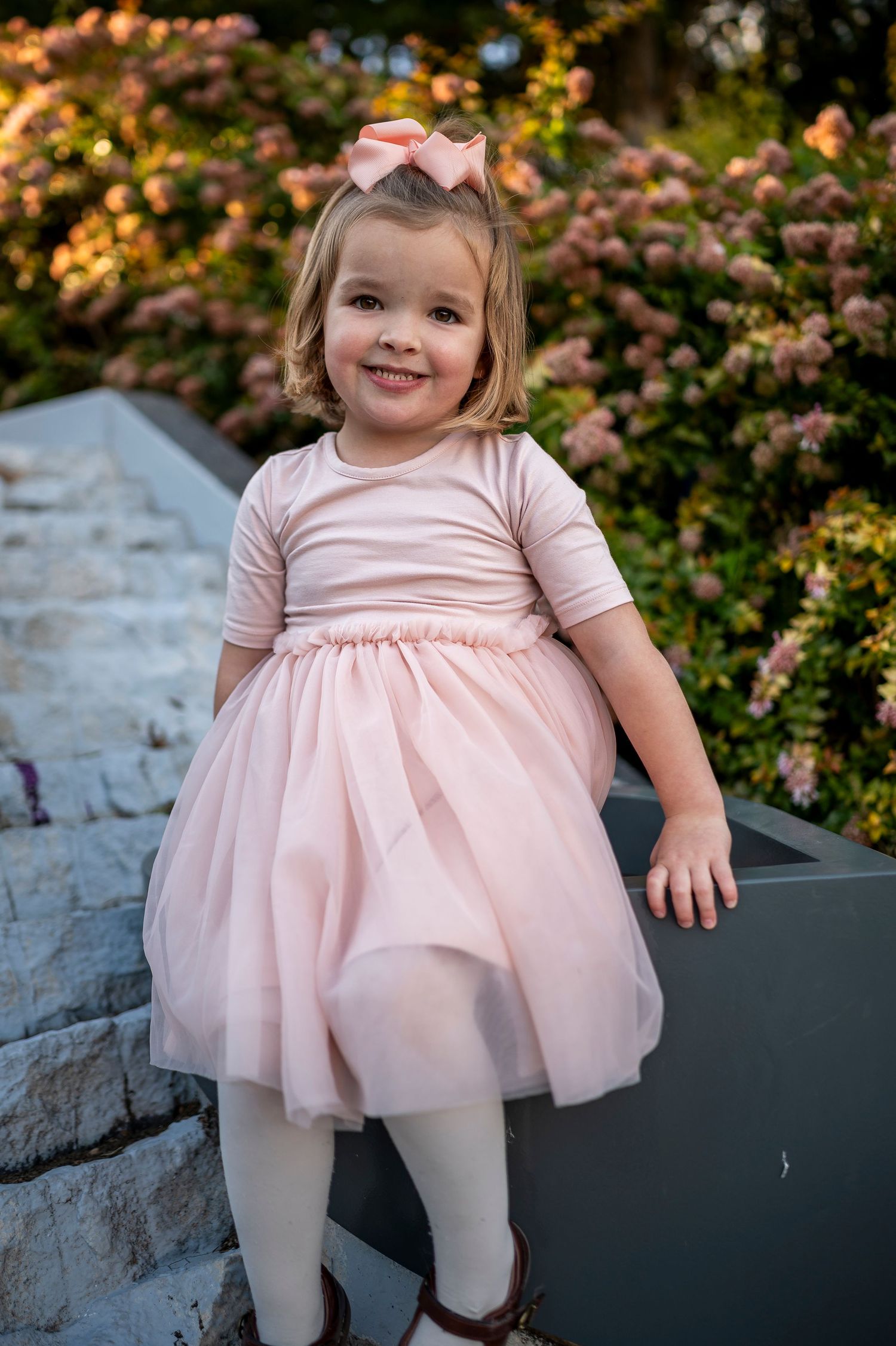 Adorable toddler girl in pink tulle dress and bow poses outdoors near blooming flowers.