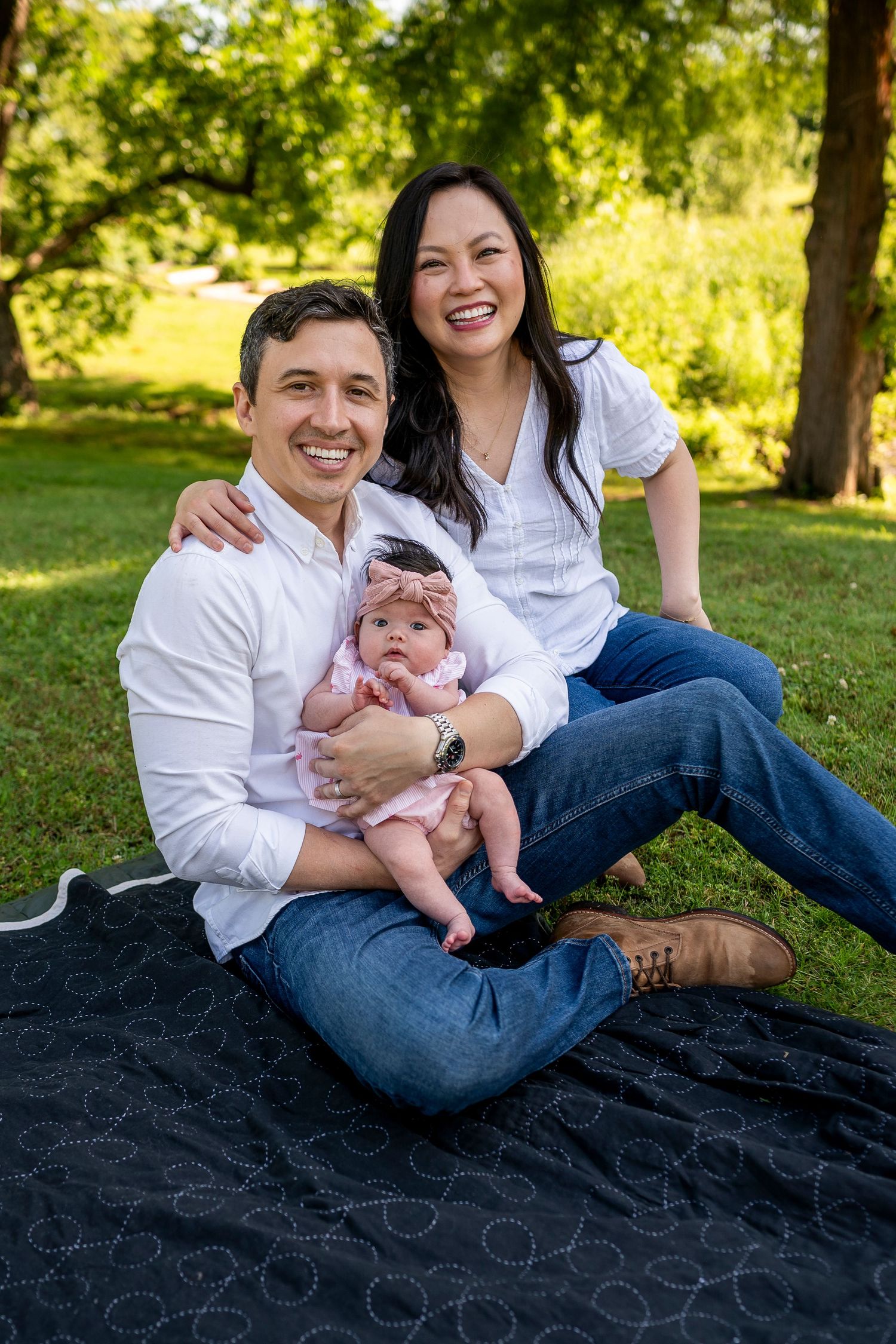 A family sits together on a black blanket in a sunny park setting, with a baby wearing a pink bow.