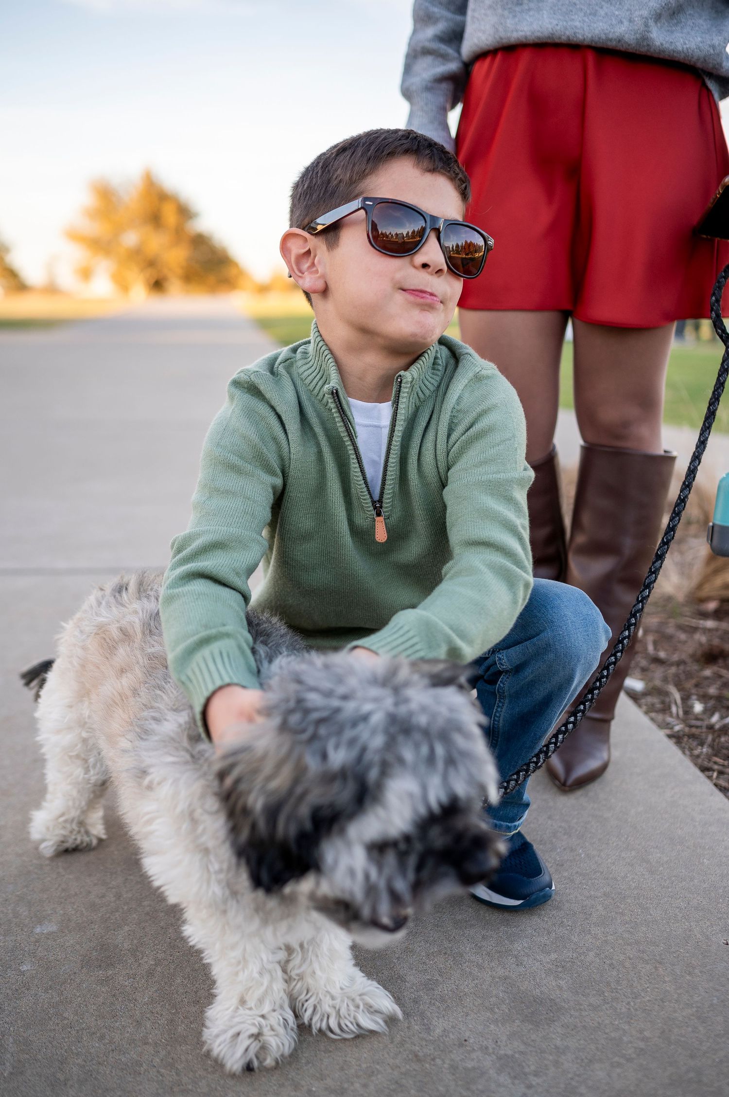 Young boy in sunglasses and green sweater crouching to pet a small fluffy dog on a sidewalk outdoors.