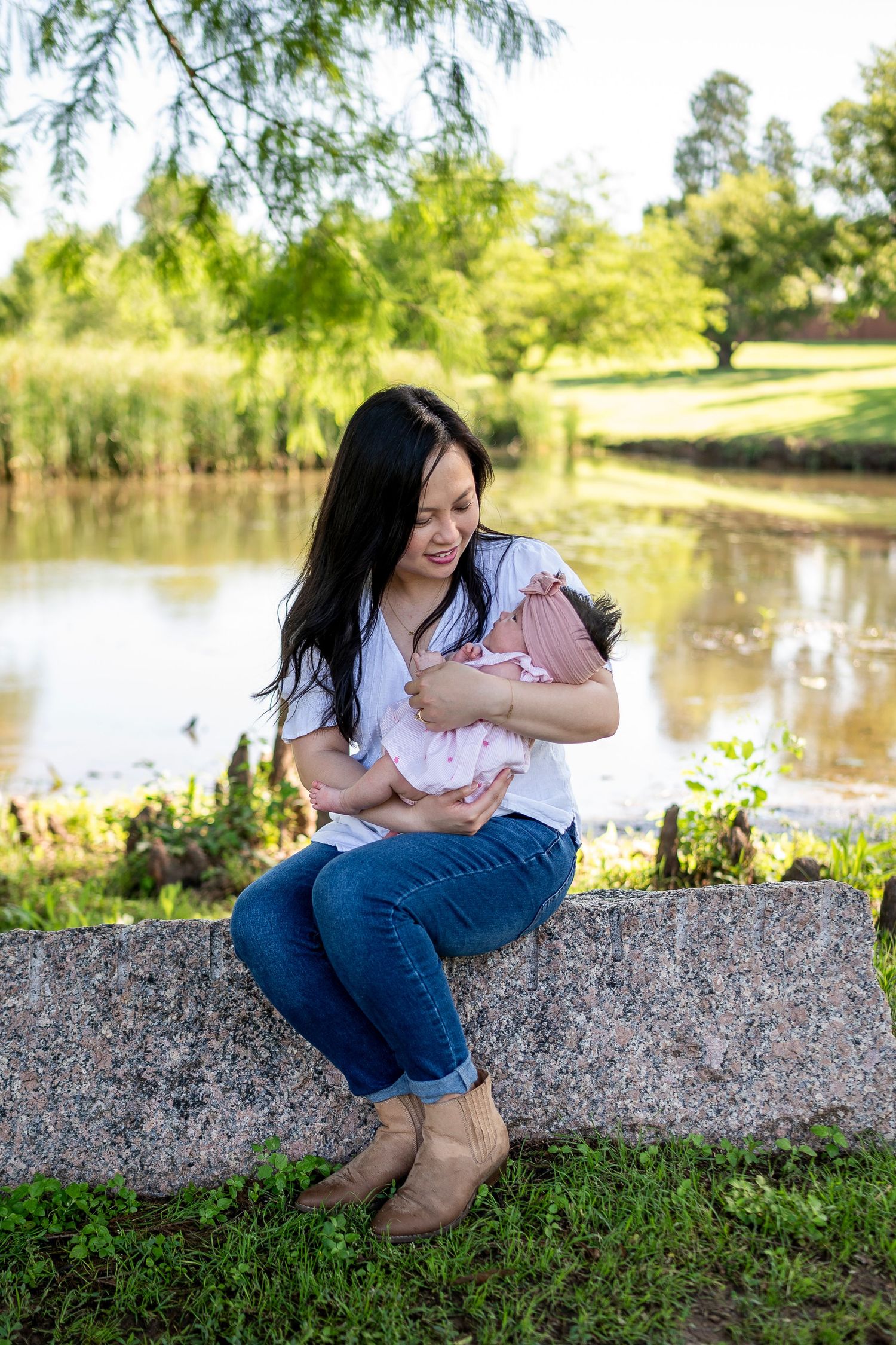 Mother and baby share a tender moment while sitting on stone wall near a serene pond on a sunny day.