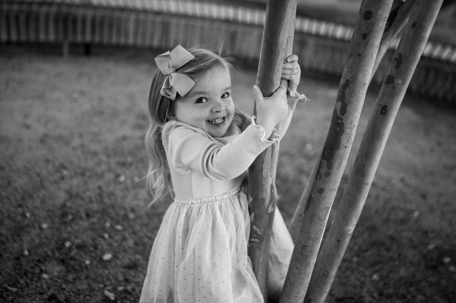 Black and white image of a small child in a dress playing on outdoor equipment.