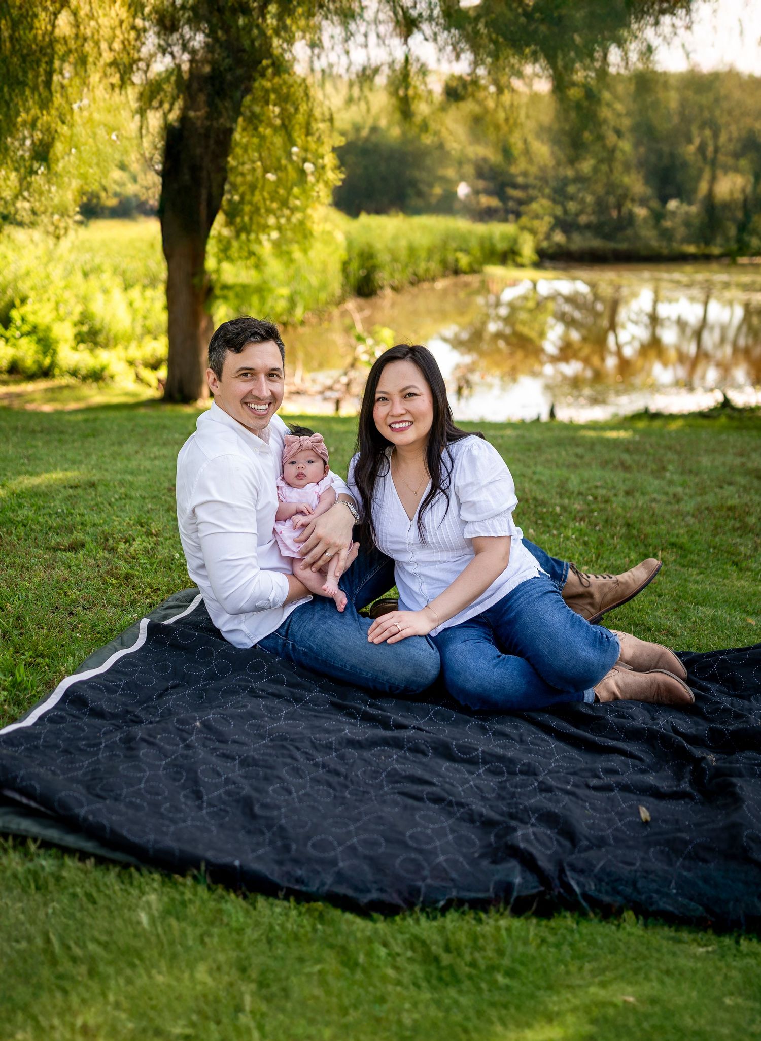 A family sits together on a black blanket near a pond on a sunny day in a green park setting.