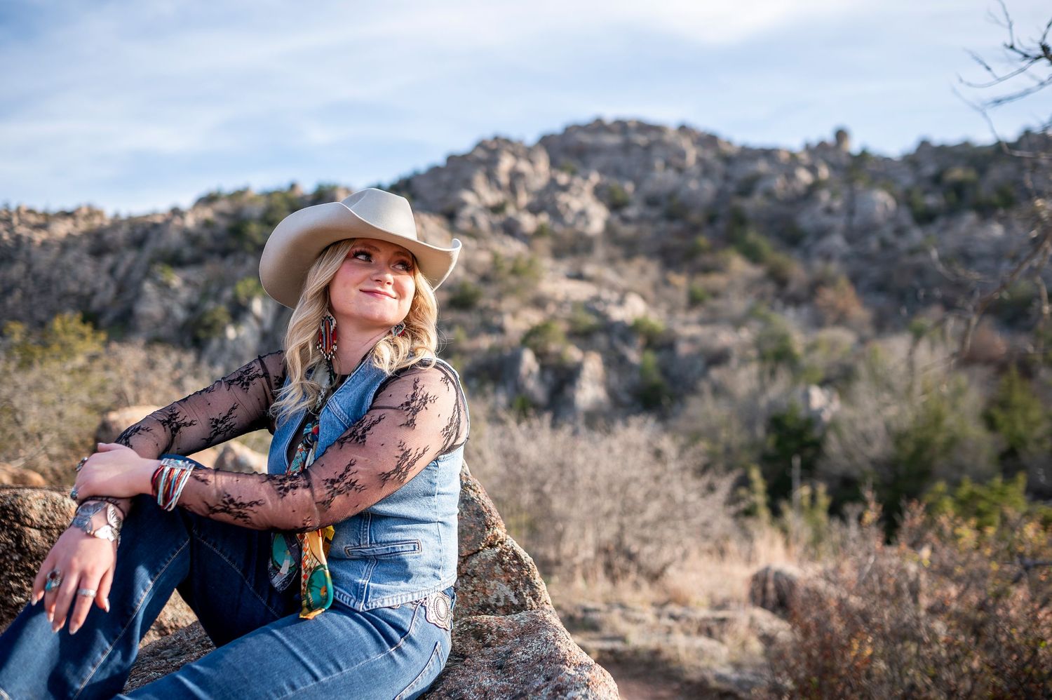 Woman in cowboy hat and jeans sitting on a rock with scenic desert hills in the background on a sunny day.