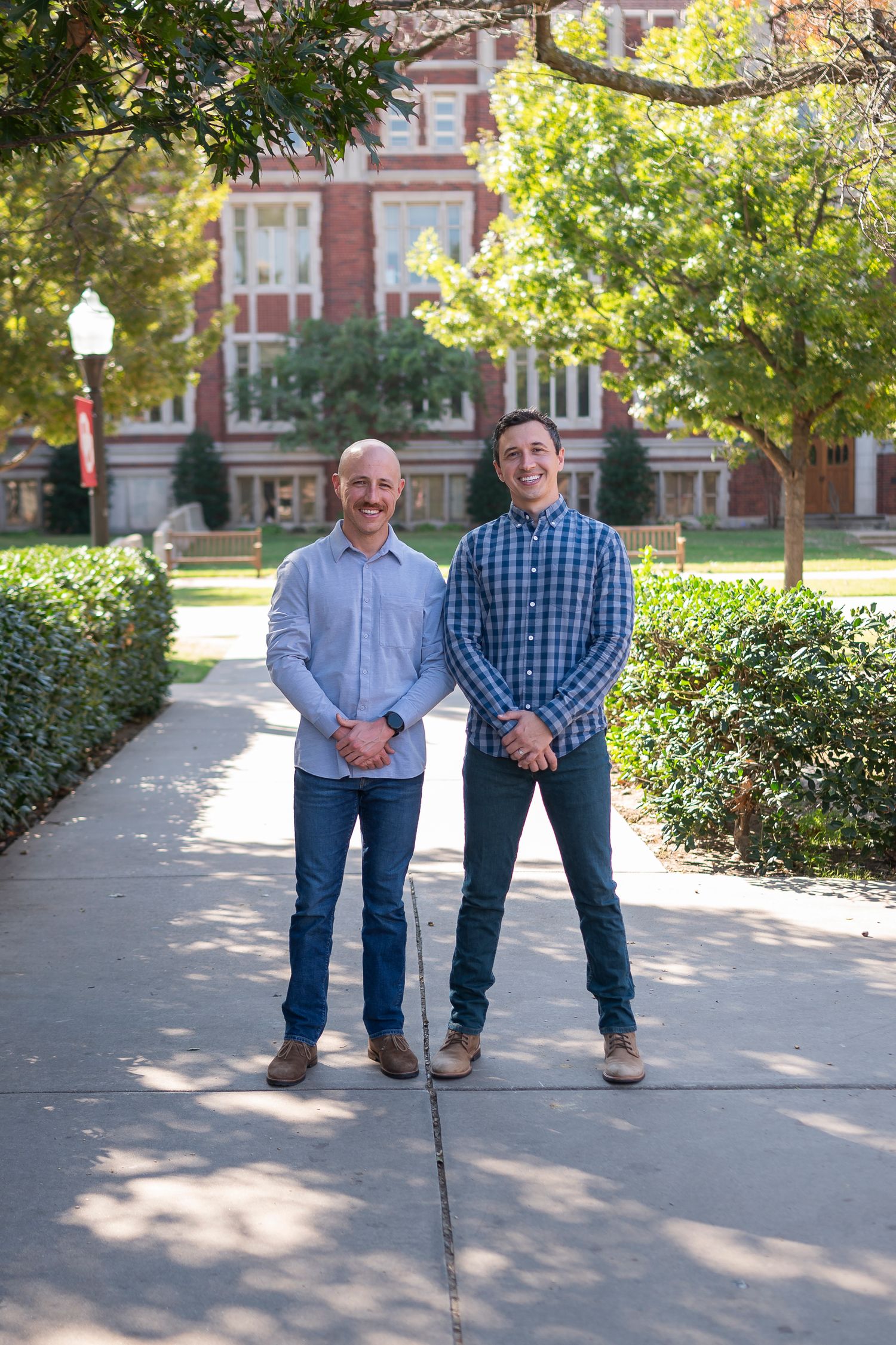 Two people standing on sunny campus walkway with red brick university building in background.