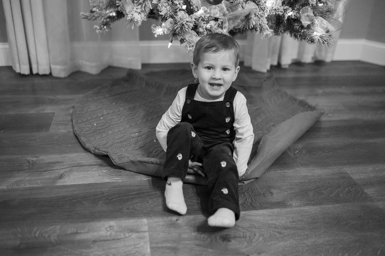 A toddler in overalls sits on hardwood flooring beneath a decorated Christmas tree.