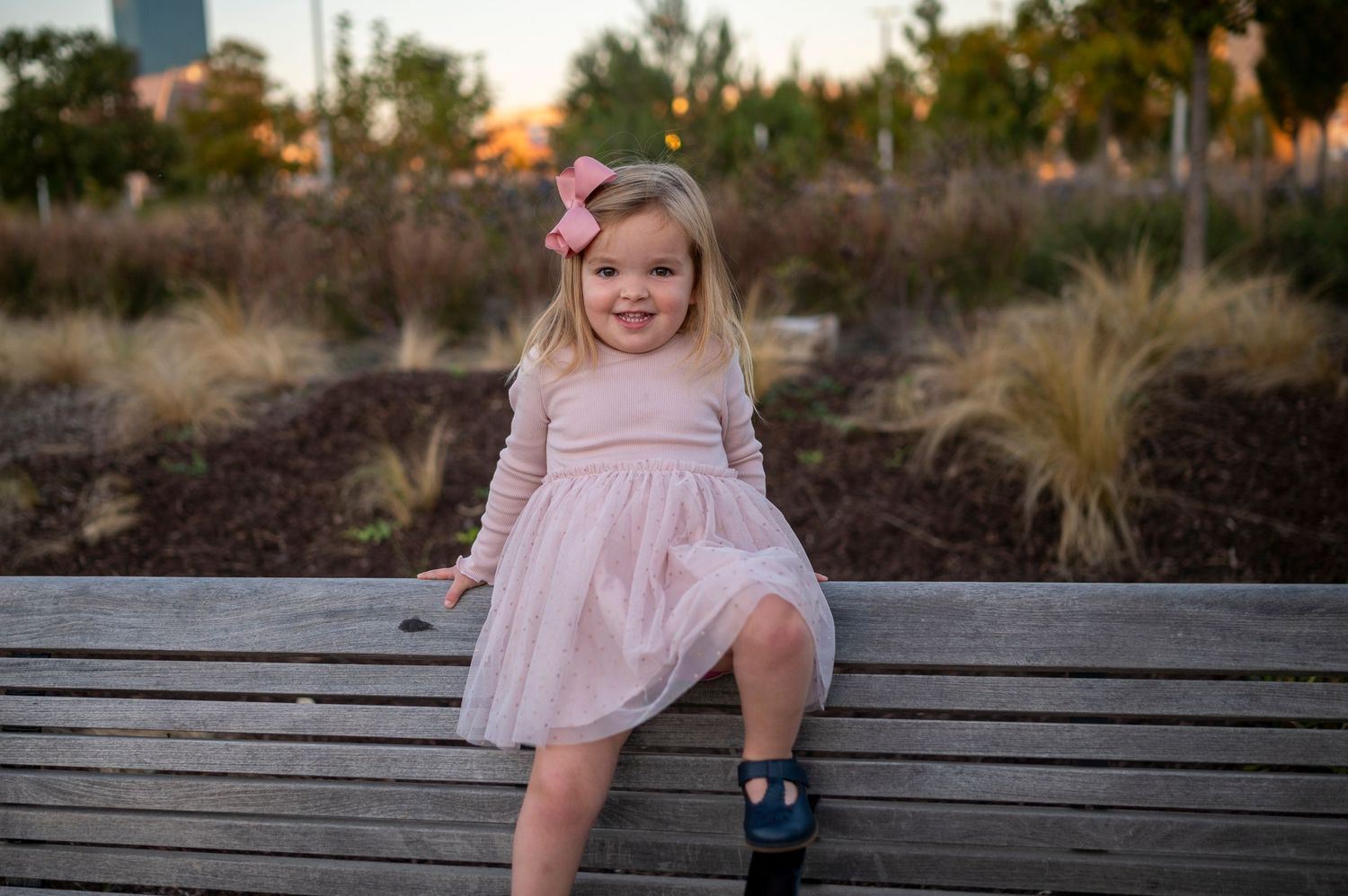 Young child in a pink dress sits casually on a wooden bench in an outdoor garden setting.
