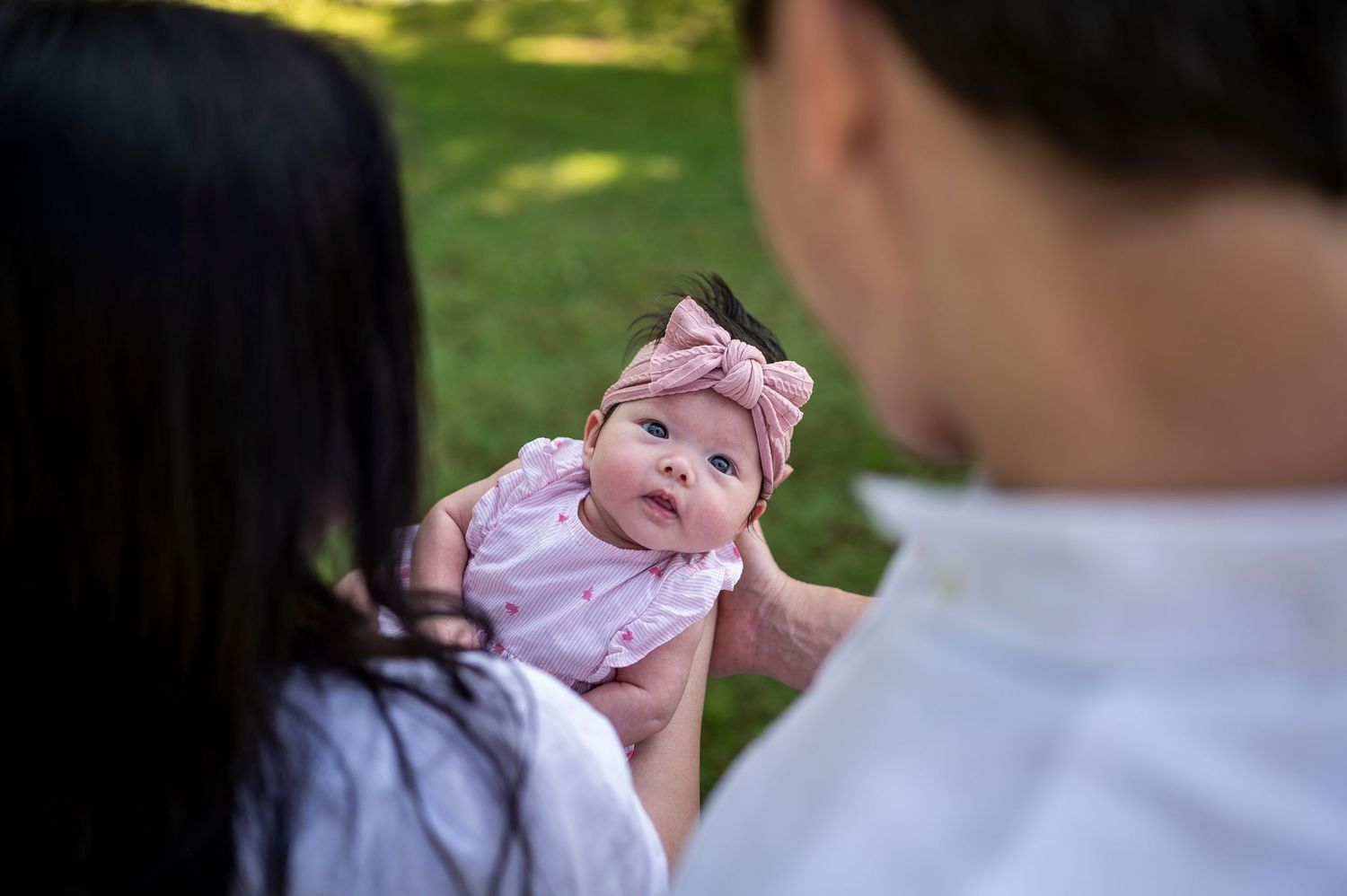 A baby in pink clothing smiles while being held up during an outdoor photoshoot.