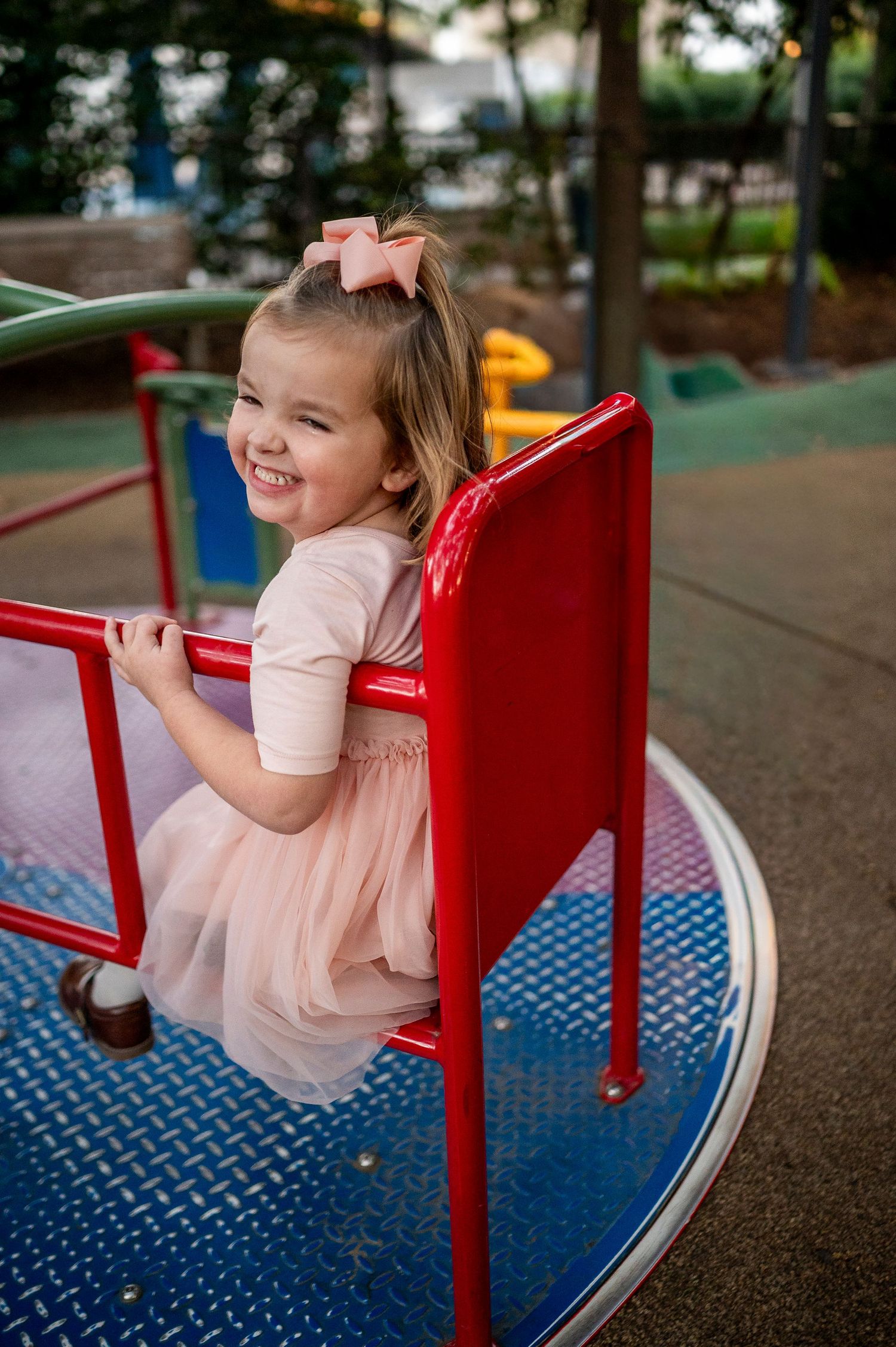 Happy young girl in pink dress riding a red merry-go-round at a playground, smiling joyfully.