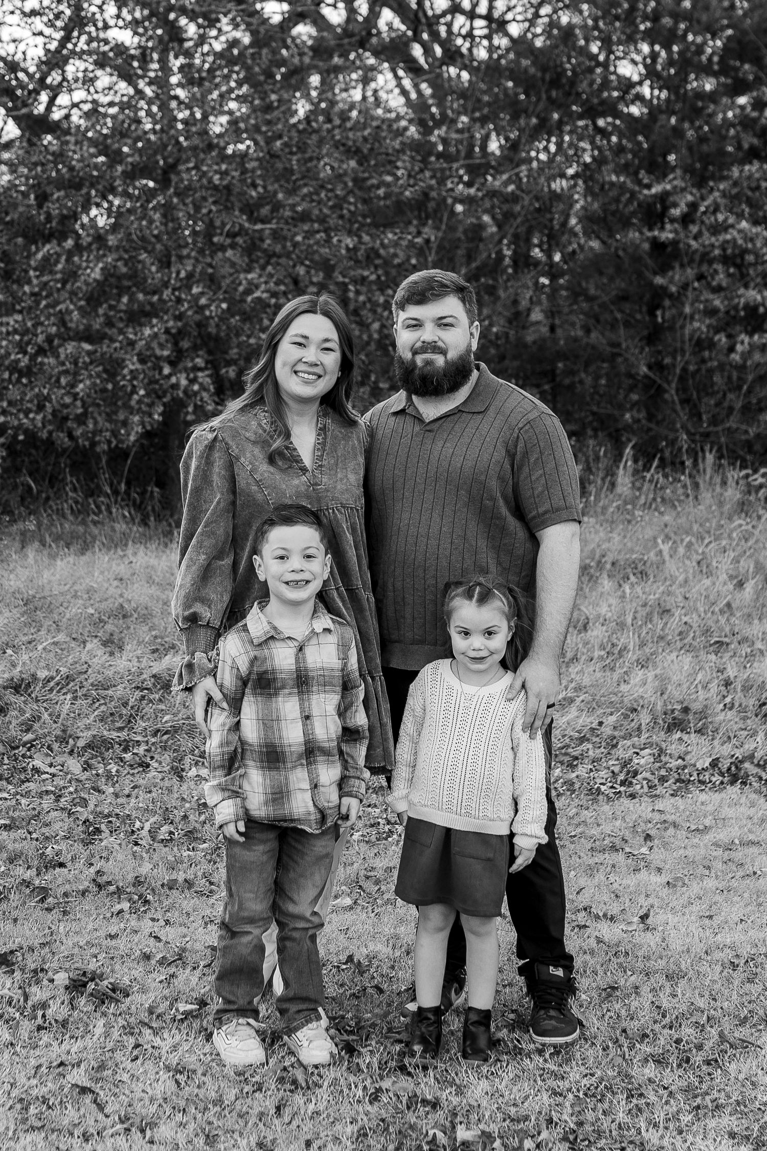 Black and white family portrait of two adults and two young children smiling outdoors in a grassy field with trees.