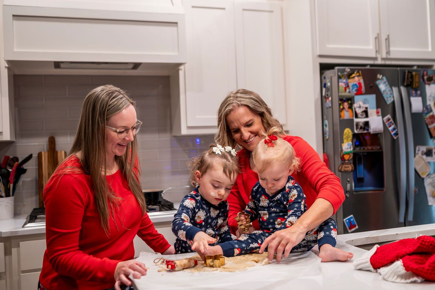 Two women in red shirts help two toddlers roll cookie dough in a festive kitchen during the holidays.