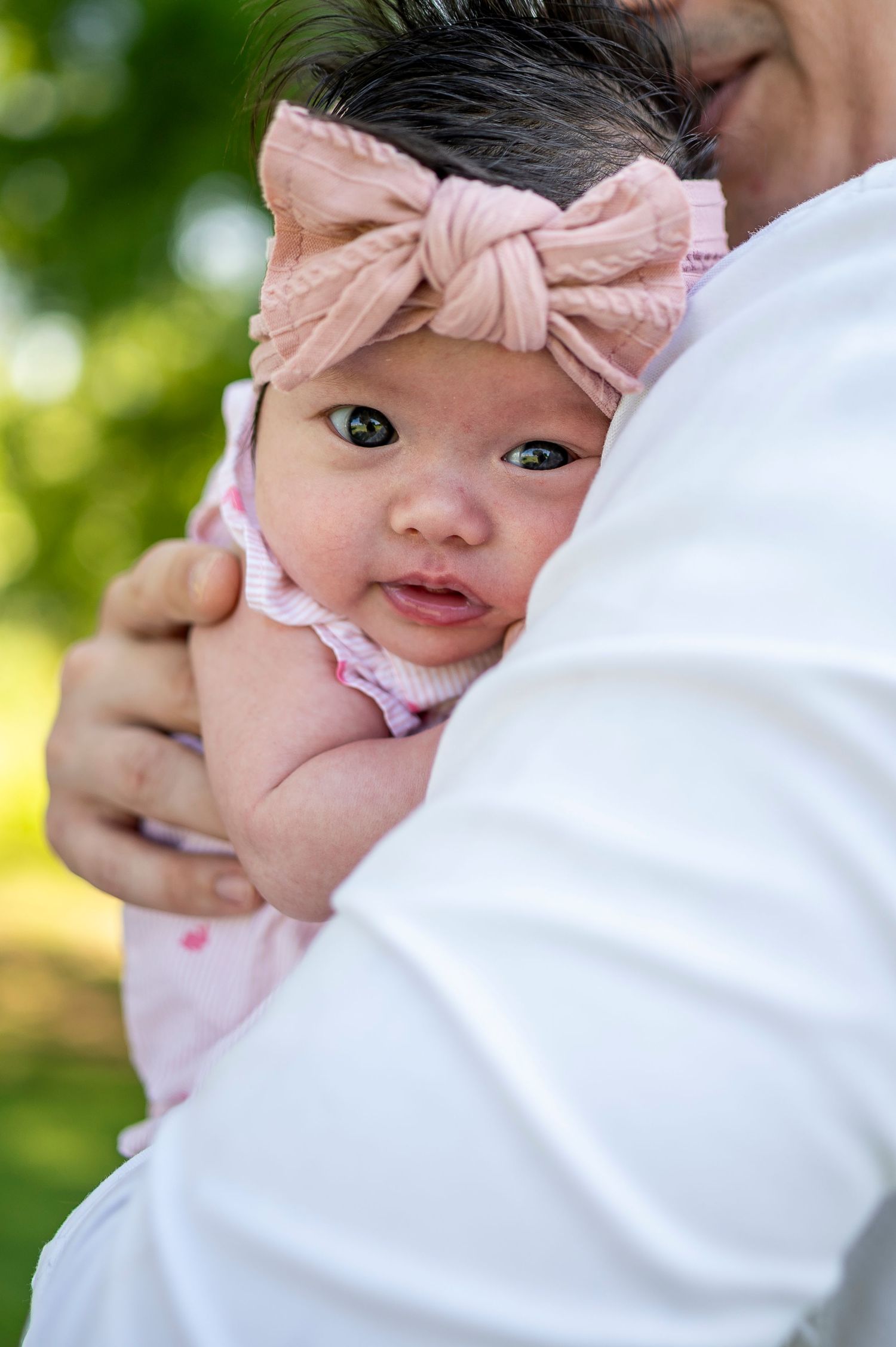 A baby in white clothing with a pink bow smiles while being held against a blurred green outdoor background.