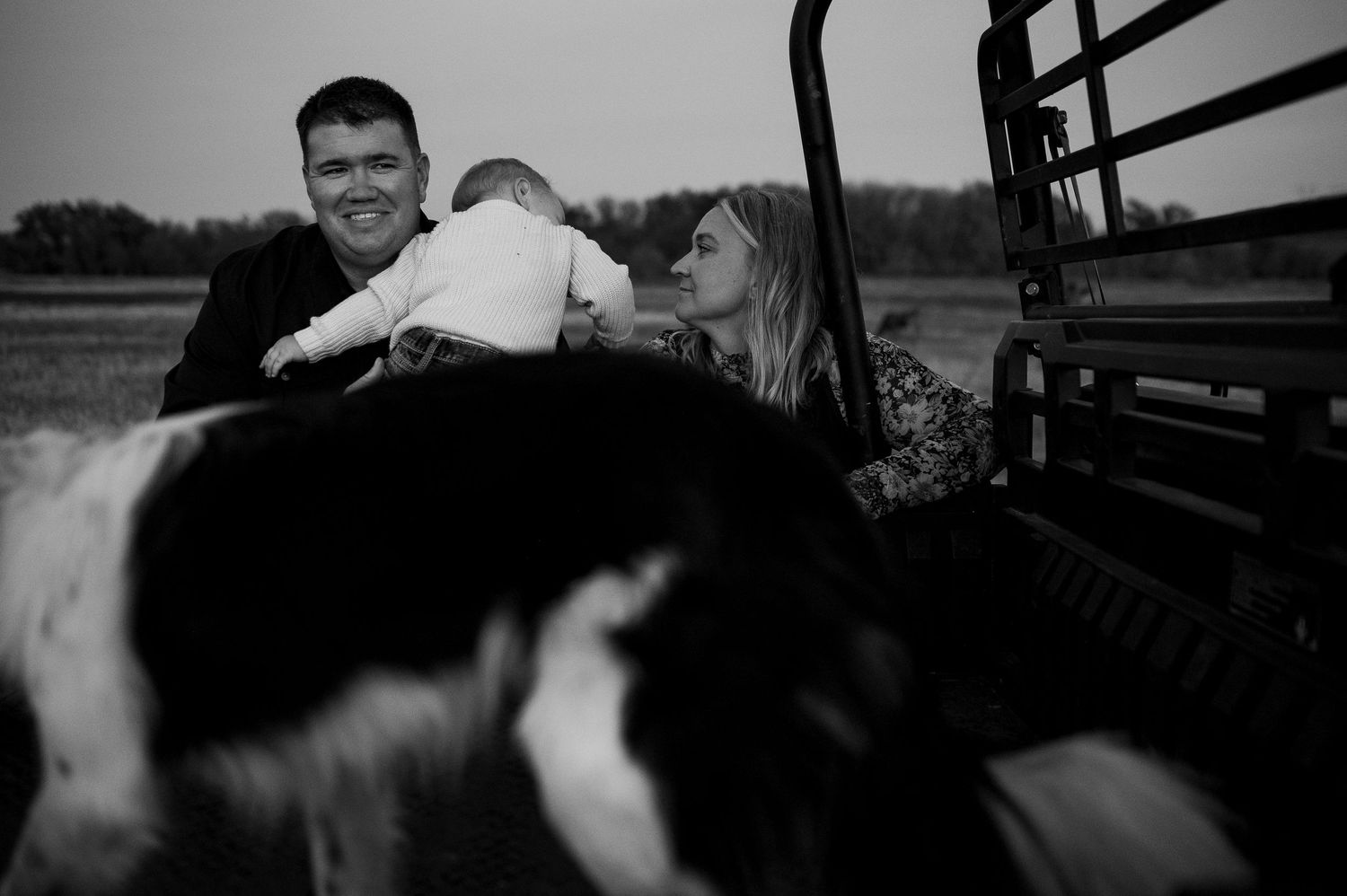 Parents and baby sharing tender moment with border collie on farm utility vehicle in black and white.