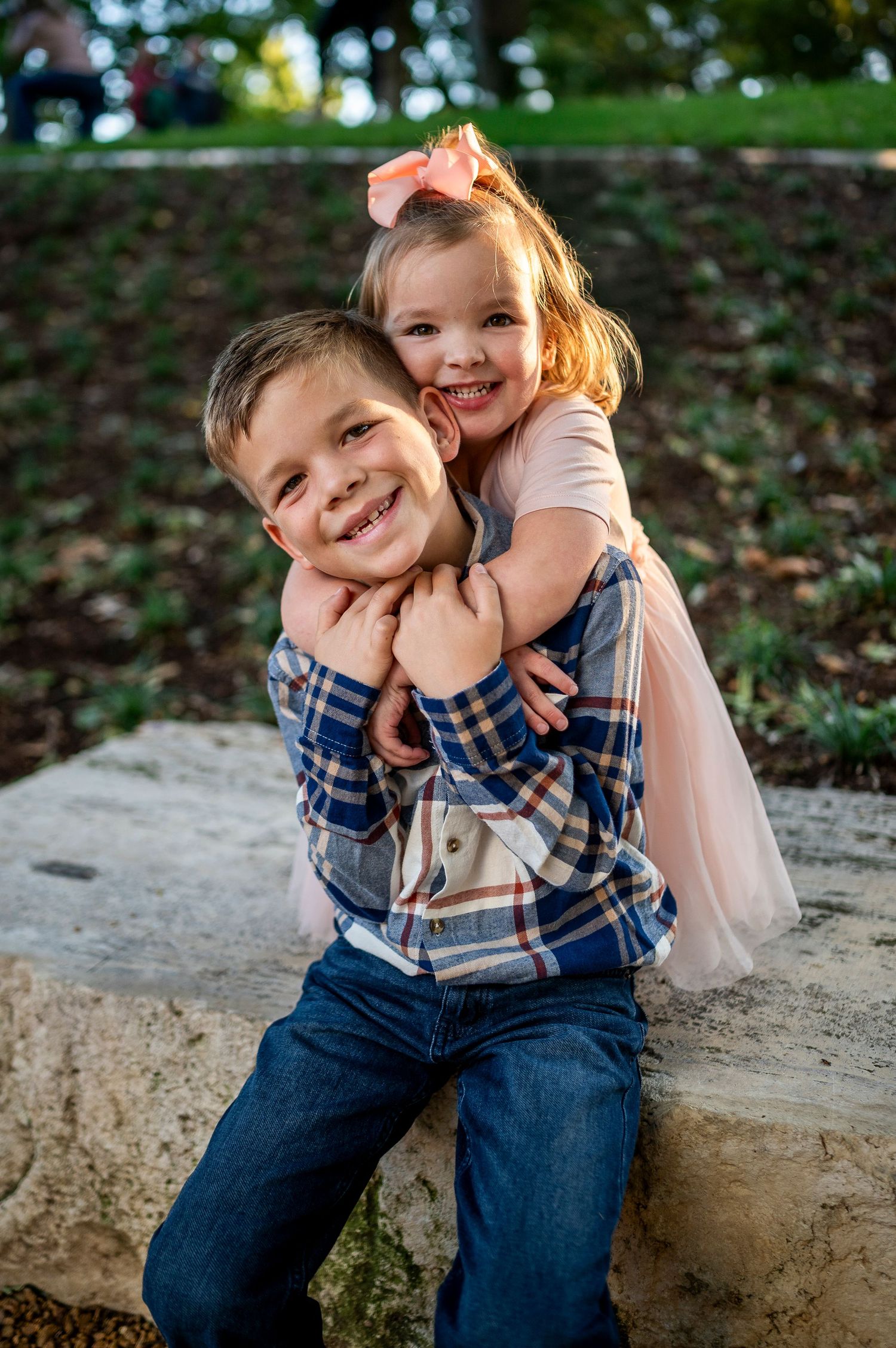 Two happy children hugging outdoors, a boy in plaid shirt and jeans with a girl in pink tutu and bow.