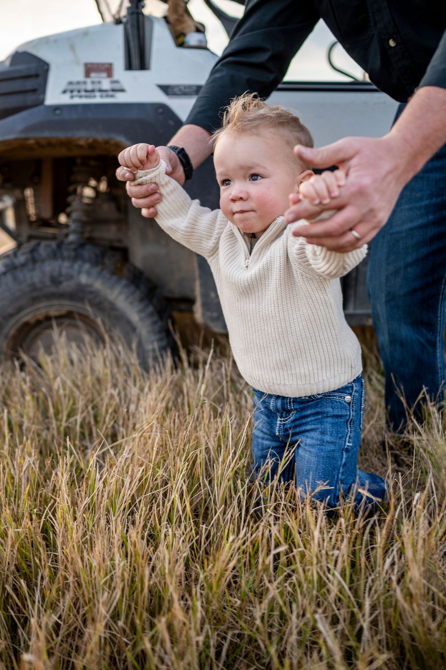 Toddler in cream sweater and jeans takes first steps in tall grass, supported by adult hands, with a tractor behind.