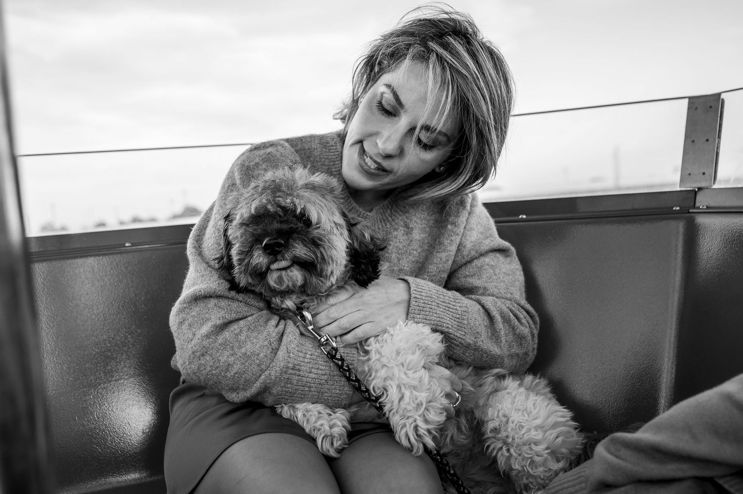 Black and white photo of a smiling woman sitting on a bench holding a small fluffy dog on her lap.