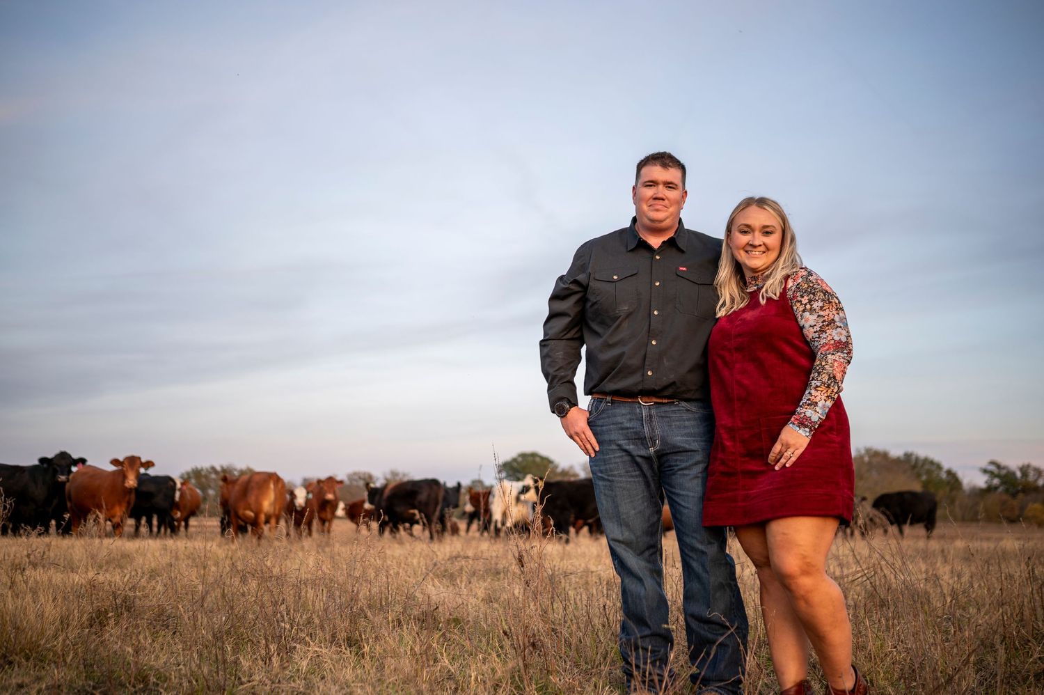 Couple smiling together in a field with cattle grazing behind them at golden hour on a ranch.