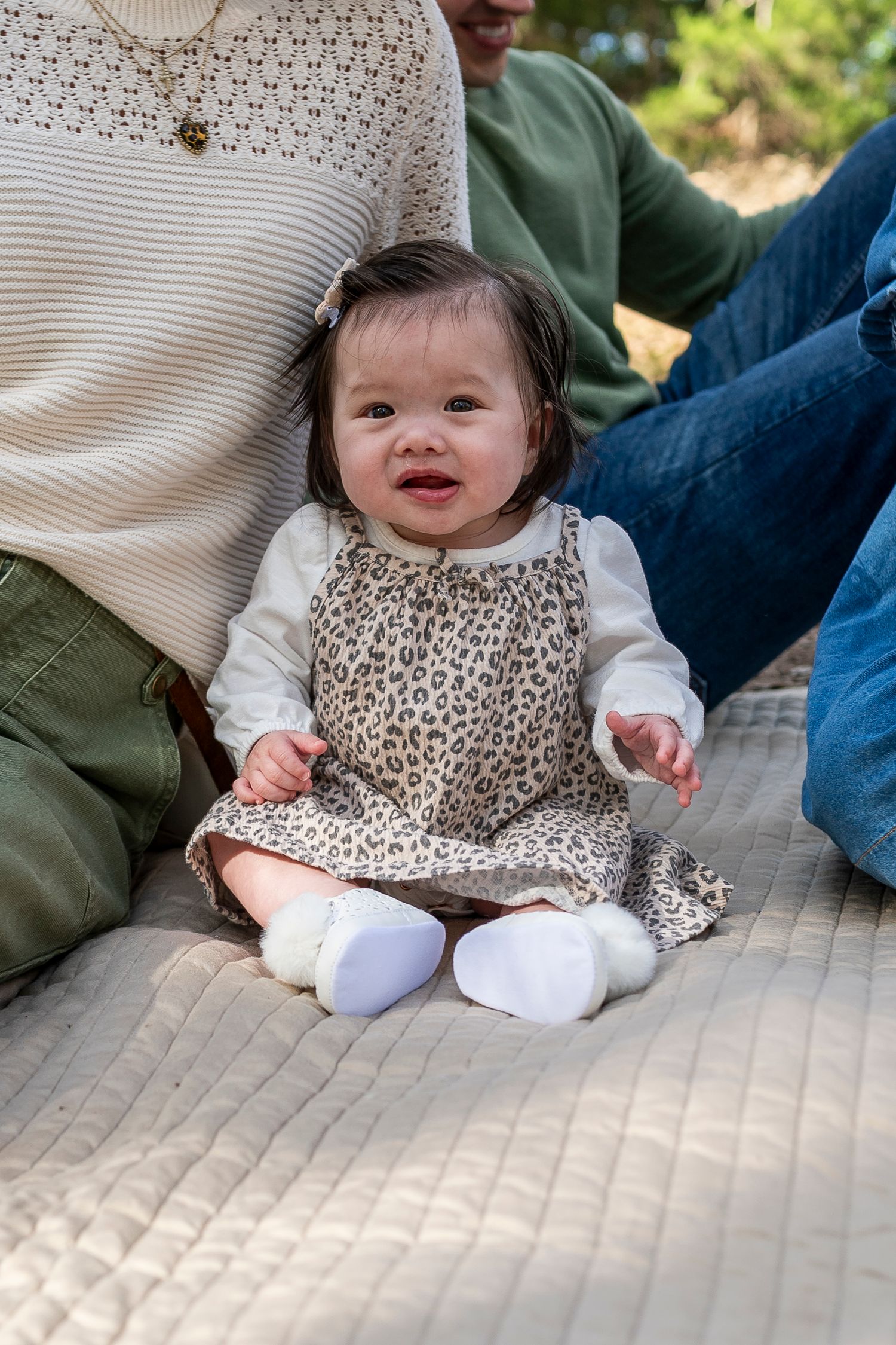 Smiling baby girl in leopard print dress and white booties sitting on a blanket between two adults outdoors.