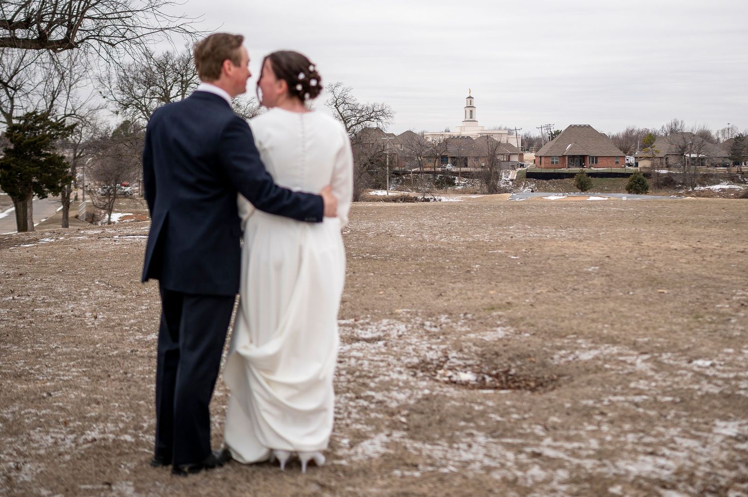 A winter wedding couple stands together overlooking a distant town landscape.