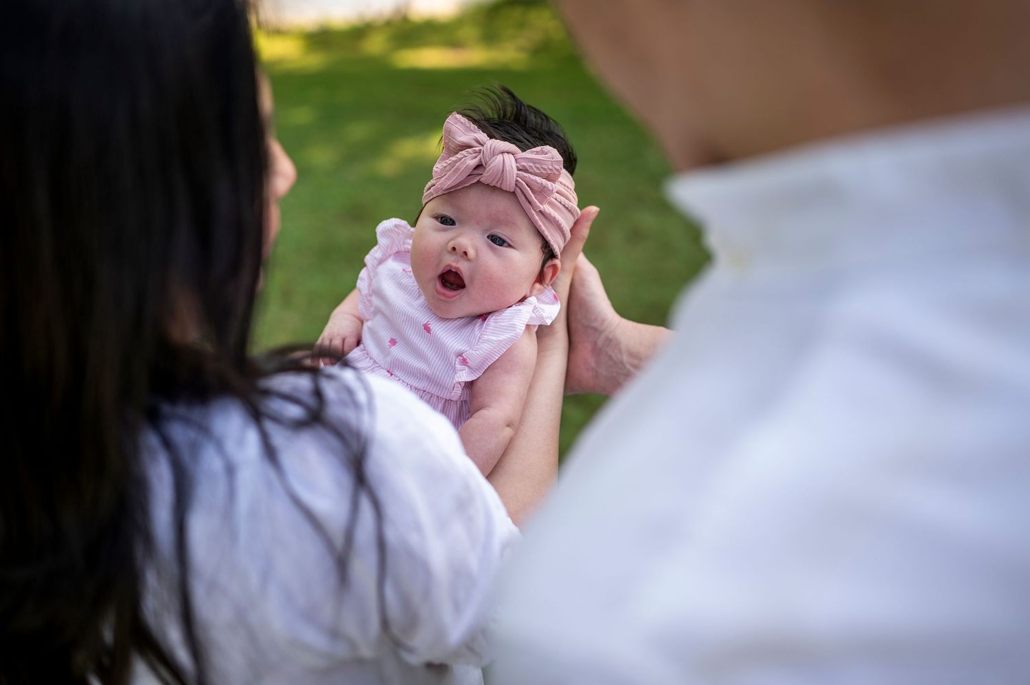 A baby wearing a pink bow headband smiles while being held up outdoors during golden hour.