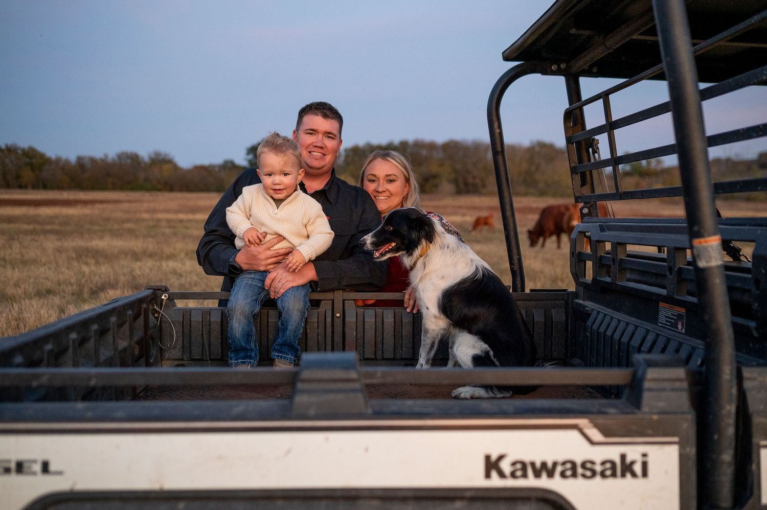 Family with baby and Border Collie dog sitting on a Kawasaki utility vehicle in a farm field.