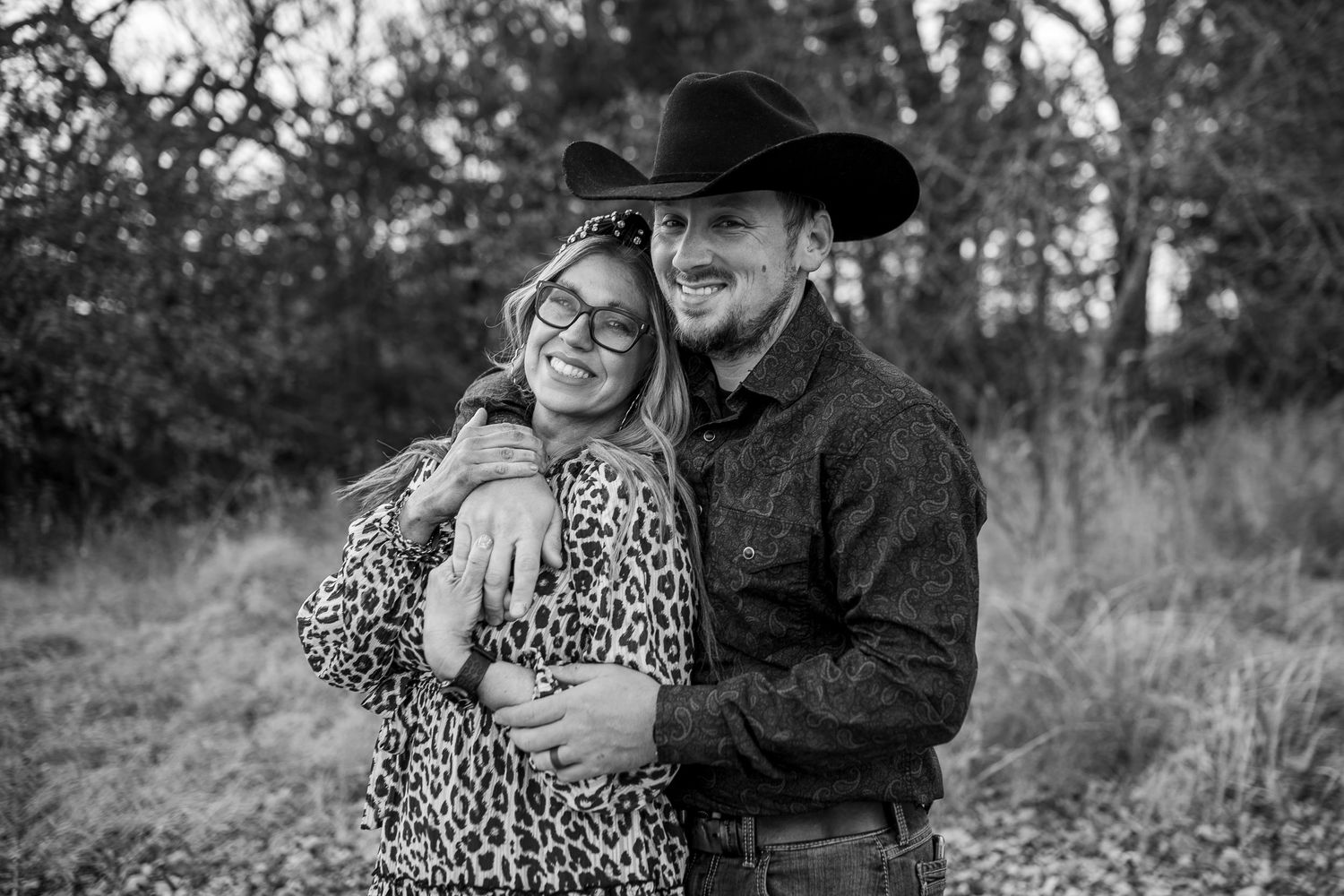 Black and white photo of a smiling couple in cowboy hats embracing outdoors among trees.