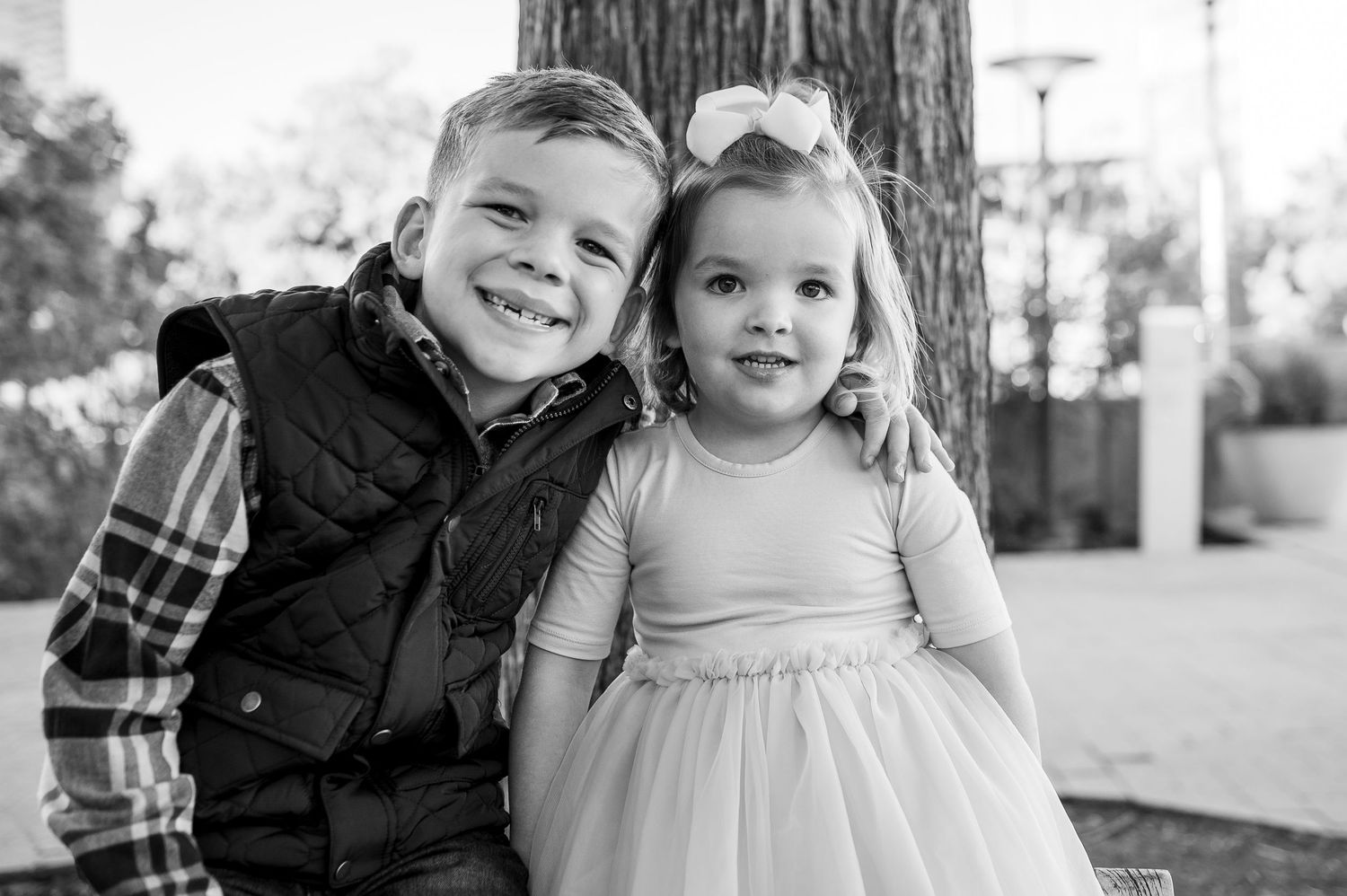 Black and white photo of two young siblings smiling together outdoors, boy in vest and girl in tutu dress.